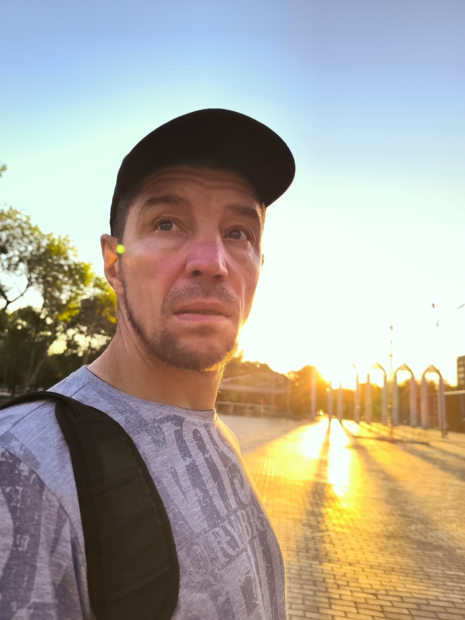 A man wearing a gray t-shirt, black baseball cap, and a backpack is seen in a close-up shot, looking to the right. The background shows a paved area with long shadows cast by the setting sun, and a row of arched structures. Trees are visible on the left. The image appears to be taken outdoors in an urban setting.