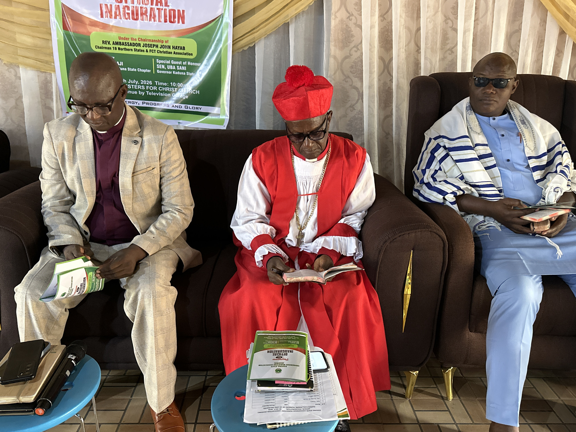 Three men are seated on a couch in what appears to be a formal event, possibly a church inauguration, as suggested by the backdrop banner. The event is stated to be in Barden Yana, Nigeria.
The man on the left is wearing a light-colored, plaid suit jacket over a deep red clerical collar shirt. He has glasses on and is looking down at a document or program he is holding in his hands. Beside him, on a small blue table, are a smartphone, a microphone, and some papers.
The man in the center is wearing a red ceremonial robe with white ruffled trim and a red headdress with a pompom. He also has glasses on and is looking down at a book he is holding open. A gold chain is visible around his neck. On the table in front of him, there are stacks of papers, a book, and a smartphone.
The man on the right is wearing a light blue traditional outfit with a white and blue striped shawl draped over his shoulders. He has sunglasses on and is looking towards the viewer. He is also holding a book.
The background banner is partially visible and features text related to an "OFFICIAL INAUGURATION" and mentions "REV. AMBASSADOR JOSEPH JOHN HAYAB" and "SEN. UBA SANI". The overall setting appears to be indoors, with patterned curtains visible in the background. The men are seated on plush, dark brown couches.