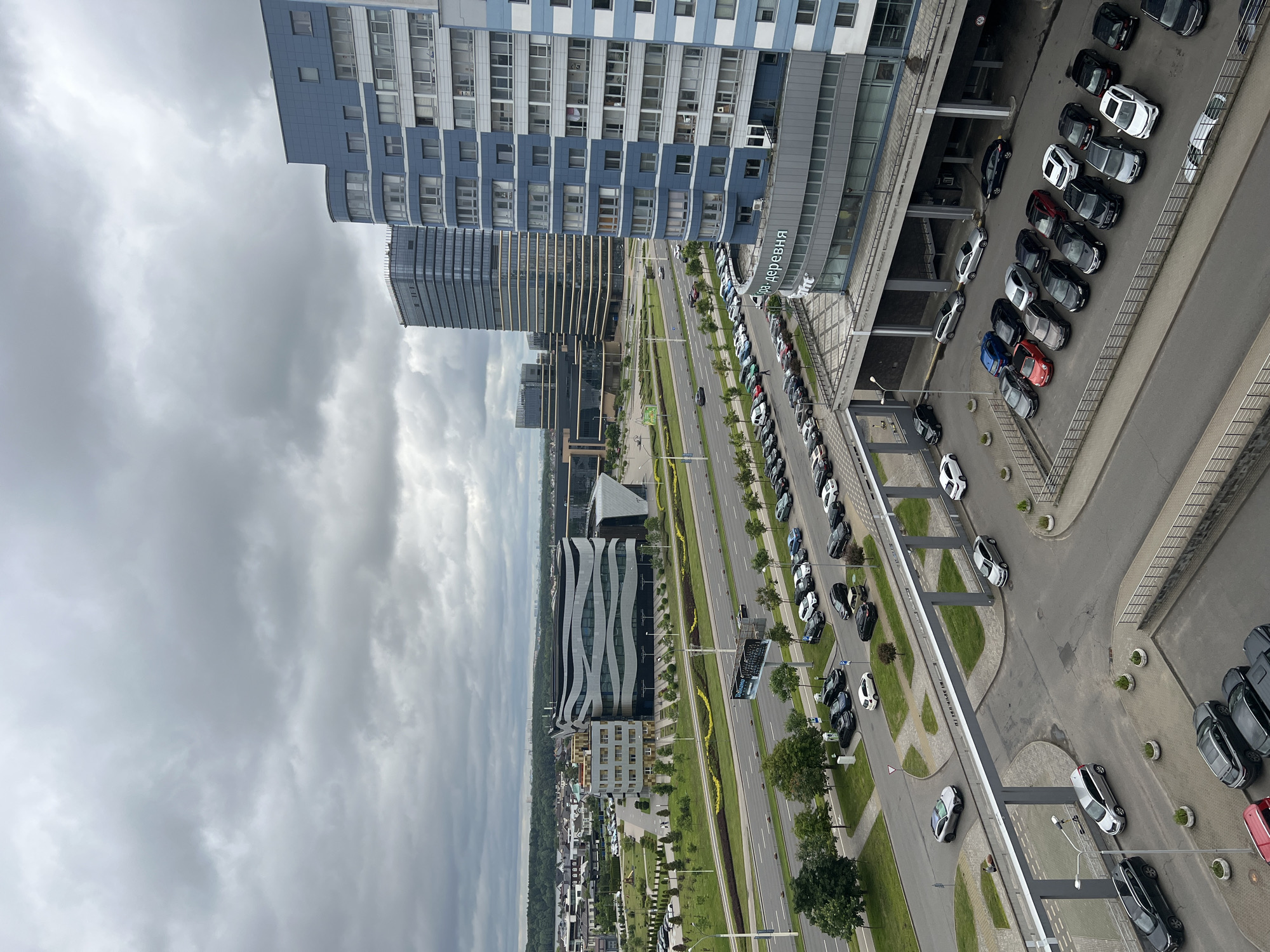 The image shows a view of a modern urban area, likely in Minsk, Belarus, under a cloudy sky. In the foreground, a multi-story blue and white residential building dominates the right side. To the left of this building, a row of parked cars is visible on a lower level, with a pedestrian walkway and railing. Further to the left and in the midground, a multi-lane road runs horizontally across the frame, with several cars in motion.
Beyond the road, a distinctively shaped modern building with a wavy facade, likely an office or public building, is prominent. Adjacent to it, taller glass-faced office buildings rise into the sky. In the background, more urban development, including residential areas and infrastructure, extends to the horizon. The environment appears to be a well-developed business and residential district. There are no visible people in the image.