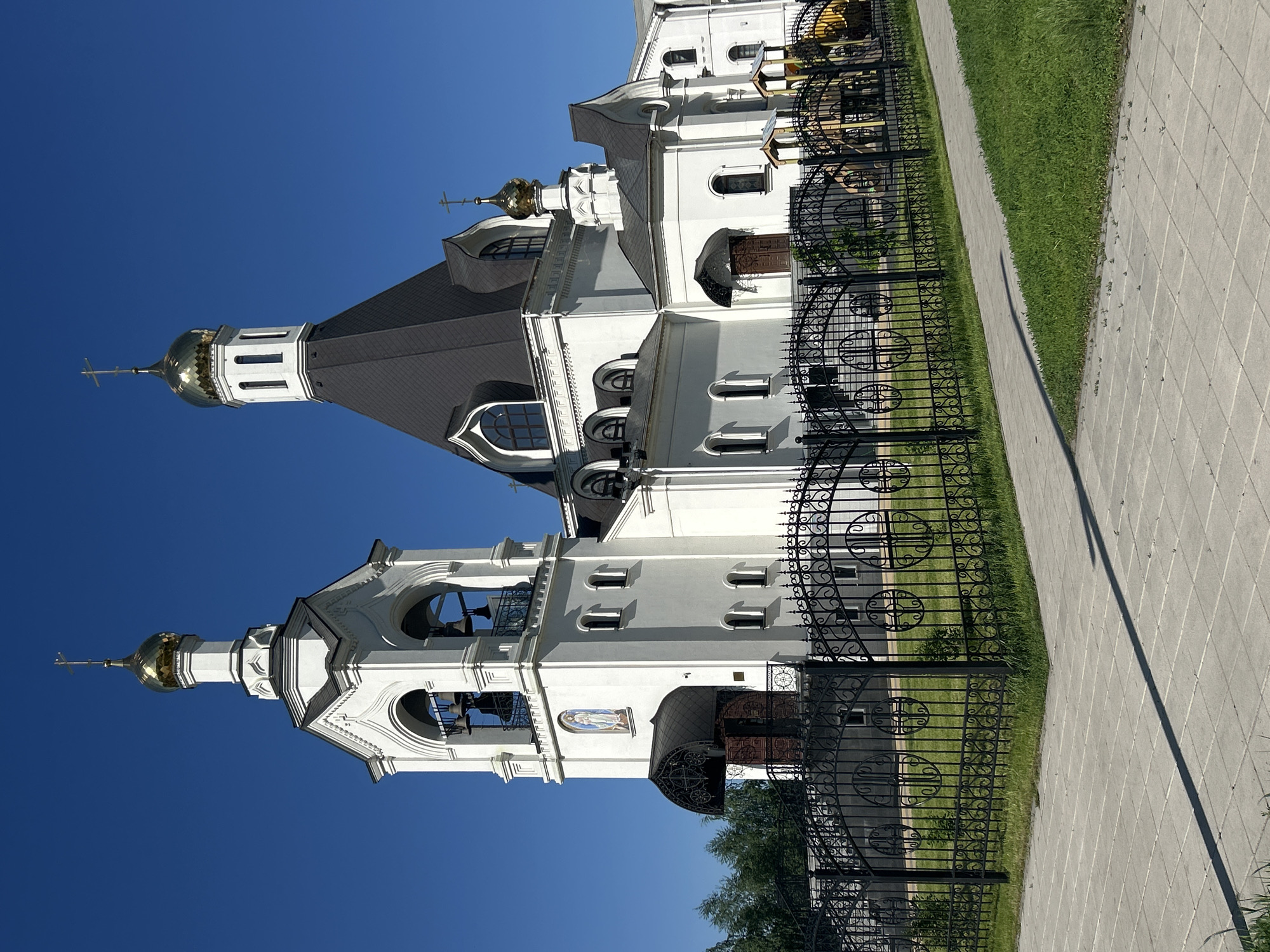 The image captures a grand, white Eastern Orthodox church complex under a bright, clear blue sky. The main subject is a large, multi-sectioned building with prominent architectural features.
The church is predominantly white, with dark grey or black sloped roofs. On the left, a tall, multi-tiered bell tower rises, topped with a gleaming golden onion dome and a cross. Bells are visible within the arched openings of the tower. Another, shorter section of the church further to the right also features a golden onion dome surmounted by a cross. The building exhibits intricate detailing, including decorative cornices, multiple arched windows, and some smaller rectangular windows. An alcove above an entrance on the bell tower section displays a religious icon or fresco, depicting a figure predominantly in blue and white. Dark, ornate wooden doors are visible at one of the entrances.
In the foreground, a decorative black wrought-iron fence with elaborate scrollwork and circular motifs runs parallel to the church. Beyond the fence, a strip of vibrant green grass borders a light grey paved walkway or sidewalk. A long, distinct shadow of the fence is cast across the walkway, indicating bright sunlight and suggesting either morning or late afternoon. The sky is a brilliant, cloudless blue, enhancing the bright and clear atmosphere. No people are visible, and no specific activity or event is taking place. There is no discernible text within the image.