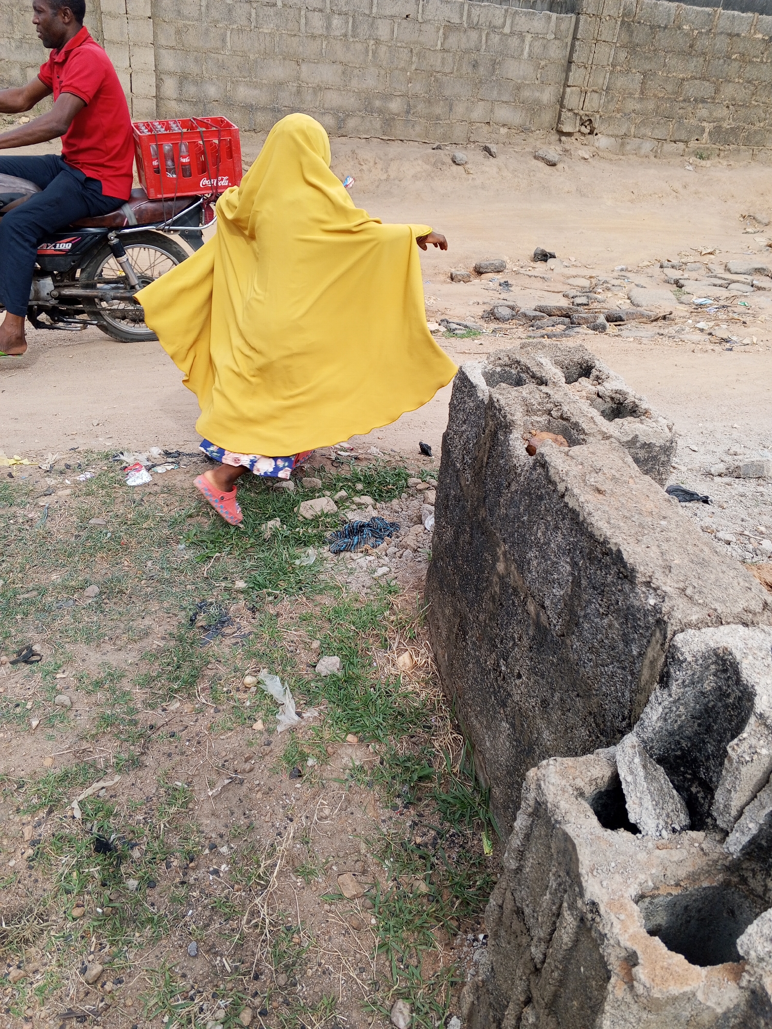 This medium shot captures an outdoor scene in Adonkolo, Nigeria, featuring two individuals and a motorcycle on a dirt path next to a rudimentary concrete wall.
On the left, a man with dark skin, wearing a red short-sleeved polo shirt and dark trousers, is seated on a motorcycle. He is shown from the chest up, in profile, looking forward. His left foot is bare, while his right foot is obscured by the motorcycle. The motorcycle, possibly a Suzuki AX100 based on visible text "AX100" on its side cover, has a red crate filled with glass bottles, clearly branded "Coca-Cola," secured to its rear rack. The man's posture suggests he is either waiting or just starting to move.
Beside the motorcycle and slightly to the right, a person, likely a child or small adult, is depicted from behind, wearing a loose, flowing yellow garment that covers their head and body. A glimpse of a patterned blue and white skirt or dress is visible beneath the yellow attire, along with a red shoe on their visible left foot, suggesting they are walking or running. Their right arm is slightly extended, as if balancing or moving dynamically.
The ground is unpaved, a mix of dirt and sparse green grass, with scattered small rocks and pieces of debris. To the far right, large, weathered concrete blocks are lying on the ground, some broken, and partially obscuring the lower right corner of the frame. In the background, a tall, rough-hewn concrete block wall stretches across the width of the image, indicating the boundary of a property or structure. The lighting suggests it is daytime with bright, natural illumination.