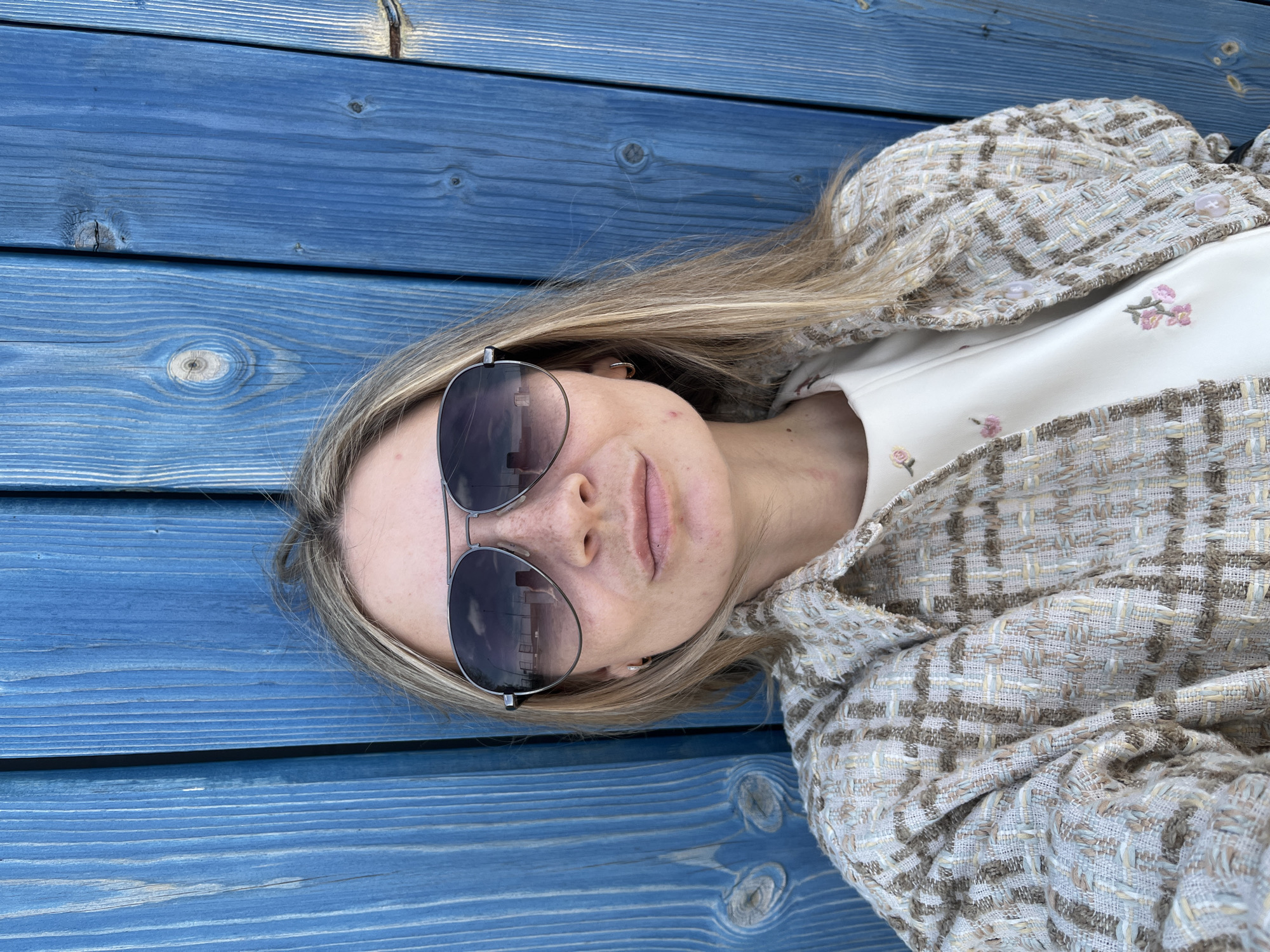 A woman wearing sunglasses and a patterned jacket is lying on a blue wooden surface, presumably a deck or platform. She is also wearing a white shirt with floral embroidery. The reflection in her sunglasses shows the presence of buildings, potentially indicating an urban environment. The wood grain of the surface is visible, and there are knots in the wood. Without further information, it is not possible to confirm if the location is Chaikovsky, Russia.