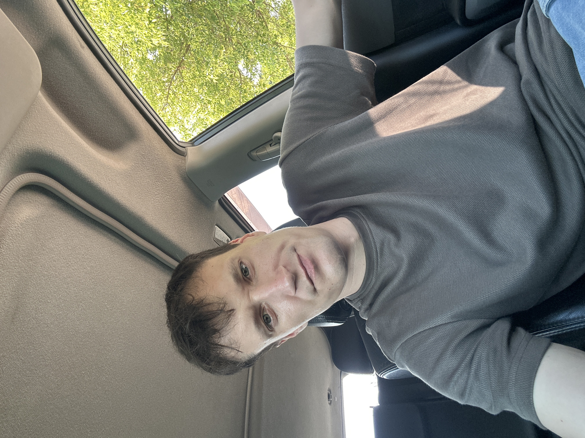 A man is sitting in the passenger seat of a car. He is wearing a grey t-shirt and blue jeans. The man is looking at the camera and smiling slightly. The car is parked outside in a sunny area with trees visible through the window. The man's head and shoulders are visible in the frame. The interior of the car is visible, including the headliner and the back seat. The lighting in the image is natural, coming from the sun.