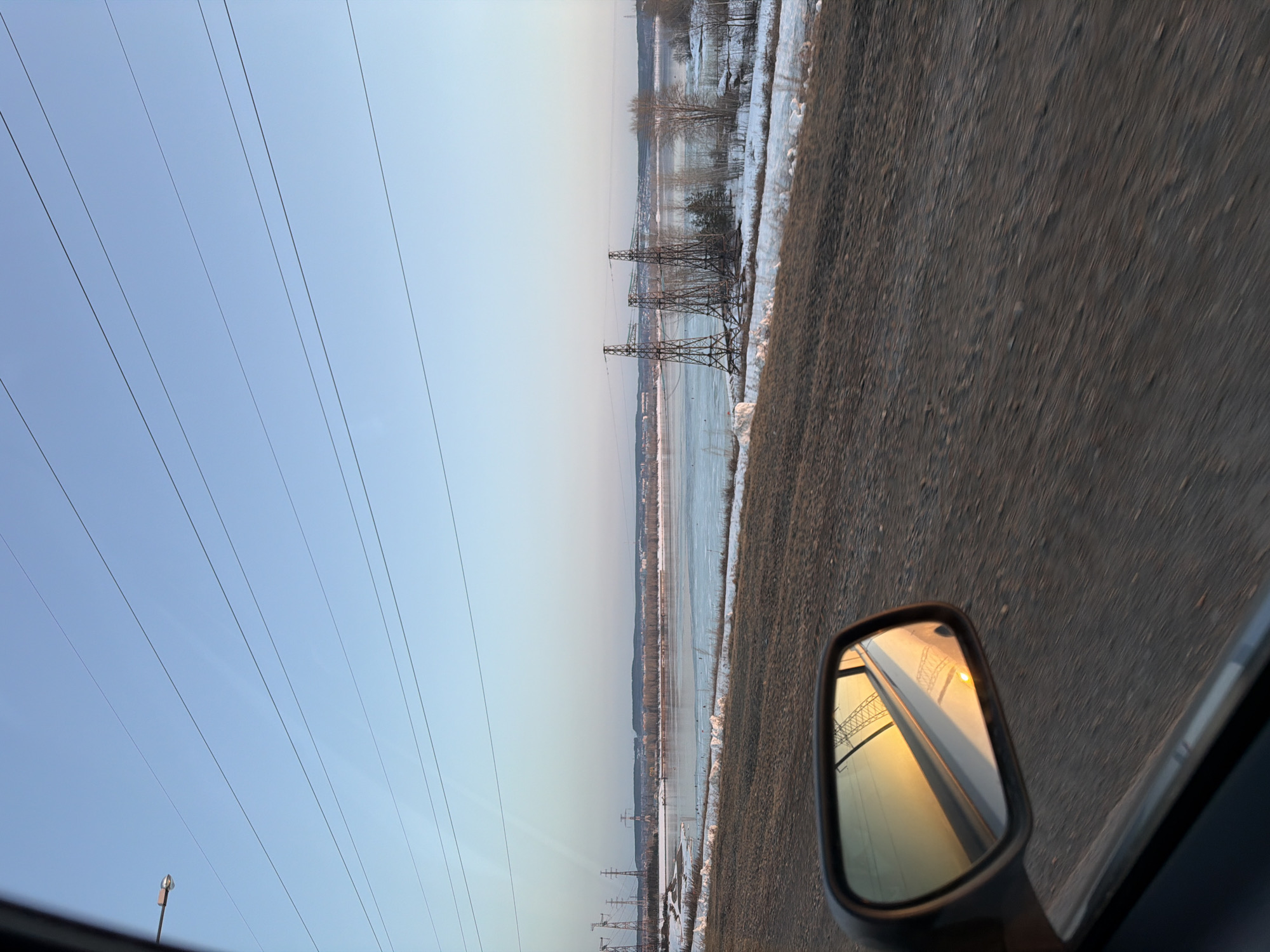 The image depicts a winter or early spring landscape in Noviy, Russia, viewed from the interior of a moving vehicle, looking outward through the passenger-side window. The scene is illuminated by the golden light of a low sun, possibly late afternoon, which is prominently reflected in the vehicle's side-view mirror in the lower-left foreground.
The sky occupies the upper two-thirds of the frame, a clear, pale blue transitioning to a deeper hue overhead. Multiple thin, dark power lines horizontally traverse the sky, and a street lamp on a pole is partially visible on the far left.
Below the horizon, a wide body of water, likely a river or large lake, dominates the midground. It is partially covered with sheets of ice interspersed with sections of open, dark water. The water's edge on the near bank features brown, patchy ground mixed with remnants of snow. Several large, lattice-structured electricity transmission towers (power pylons) stand along this bank and extend into the distant landscape. Leafless trees and shrubs are scattered along the water's edge and on a distant landmass across the water.
In the far background, a low-lying range of hills or land rises, topped with a dense line of bare trees or forest. A hazy outline of a distant town or city with various structures is visible across the water.
The immediate foreground consists of a dark, textured dirt or gravel surface, blurred horizontally, indicating the vehicle's motion. The side-view mirror reflects the golden light, a section of the vehicle's body, and a distorted image of a power pylon. No people are visible in the scene.