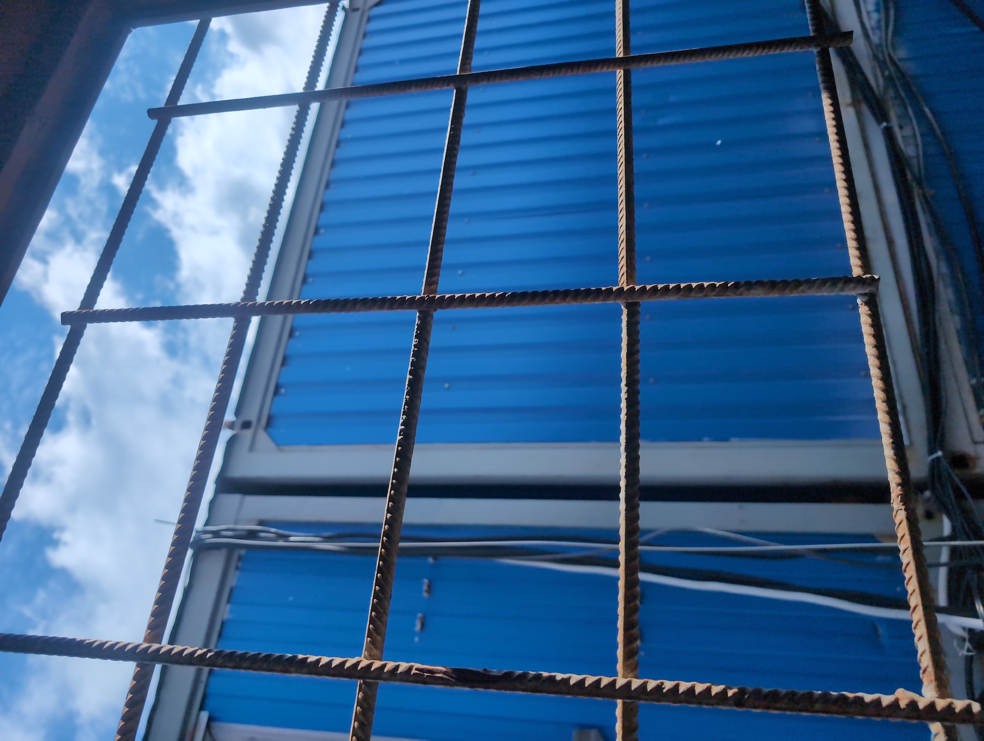 The image shows a close-up view of rusty rebar forming a grid pattern in the foreground. Through the rebar, a bright blue, corrugated metal wall is visible, likely part of a building or structure. Above the blue wall, a clear sky with white, puffy clouds can be seen. The rebar casts shadows on the blue wall, suggesting it is daytime. Wires and cables are also visible on the right side of the image, attached to the structure. The overall impression is one of an unfinished or industrial construction site. There is no visible text.
