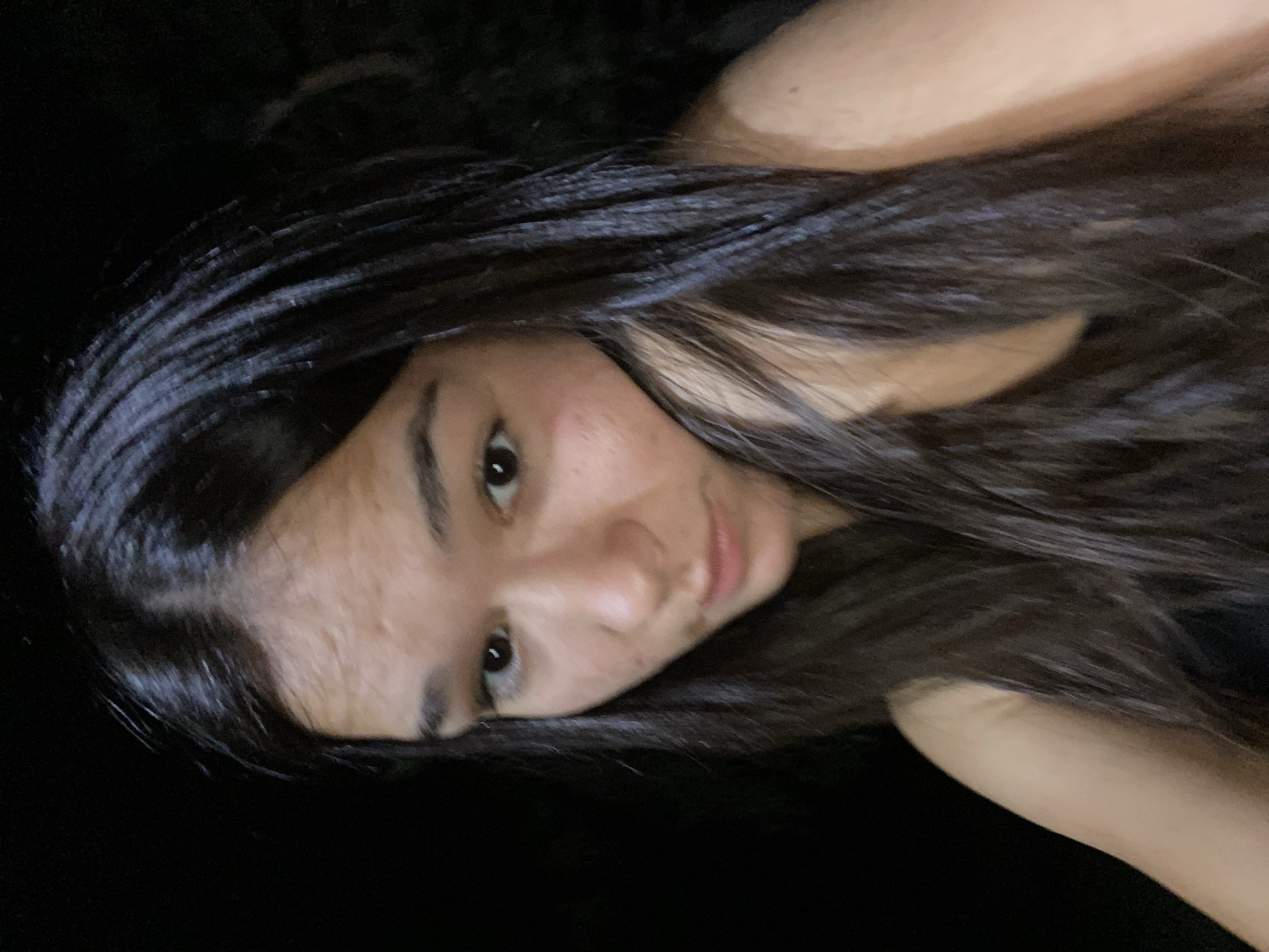This is a close-up, low-angle, indoor selfie of a young woman with long, dark brown hair. She is looking down and to her left, with her left cheek resting on her hand and her arm. The lighting is dim, with the background appearing mostly black. She is wearing a dark-colored, sleeveless top. The image appears to be taken at night. The location in the city of Hirevo, Bulgaria is not discernible from the image.