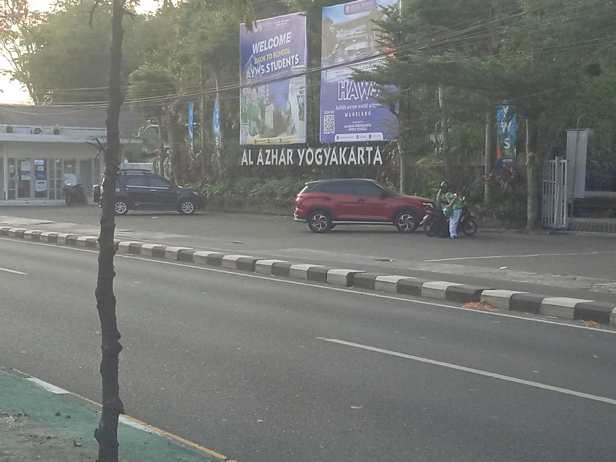 This is an eye-level, outdoor, medium shot taken in Ngaglik, Indonesia, featuring a street scene with parked cars and signage. On the left side of the image, a dark-colored SUV is parked in front of a building that appears to be an educational institution. To the right of the SUV, a red SUV is parked. A motorcycle with two individuals wearing green is also parked nearby. Dominating the background are several large banners and signs, the most prominent displaying "AL AZHAR YOGYAKARTA" in large, dark lettering. Above this, two large purple banners are visible: one reads "WELCOME BACK TO SCHOOL AYWS STUDENTS," and the other shows "HAWS hafidh asrom world sch MAIGELANG." The environment is characterized by trees, overhead wires, and a paved road with a distinctly marked curb. The lighting suggests it is daytime, possibly morning or late afternoon due to the soft quality of light and shadows.