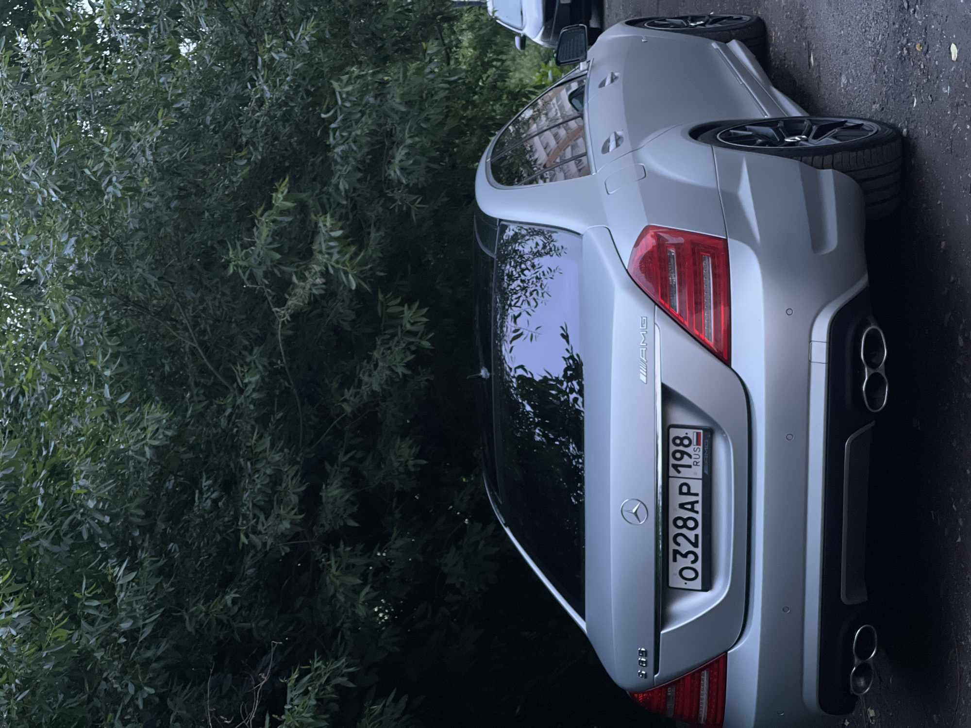 An eye-level, medium shot shows the rear passenger side of a silver Mercedes-Benz S-Class sedan parked on a paved area next to a large, leafy green tree. The car's license plate, which reads "O328AP 198 RUS", is clearly visible. The car's taillights are illuminated, and its tinted windows reflect the surrounding environment, including buildings. The car has black rims and quad exhaust pipes. A portion of another car is visible to the right of the Mercedes. The scene appears to be outdoors, possibly in a residential or urban area, with the dense foliage of the tree dominating the upper portion of the frame.