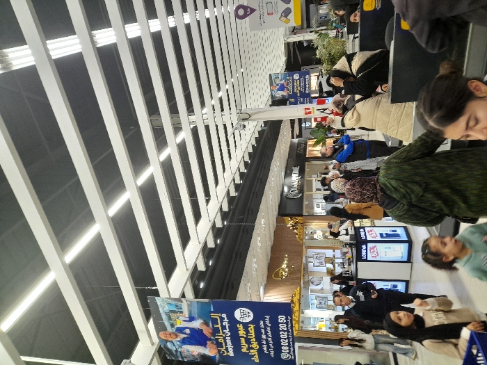 The image shows an indoor scene, likely a shopping mall or airport terminal, with a high ceiling featuring parallel white beams. A variety of shops and people are visible in the background. Prominent signage includes an advertisement for "Marjane," an establishment offering express checkout services. Text in Arabic is present on the signage, indicating a region where Arabic is spoken. The scene suggests a busy commercial environment.
