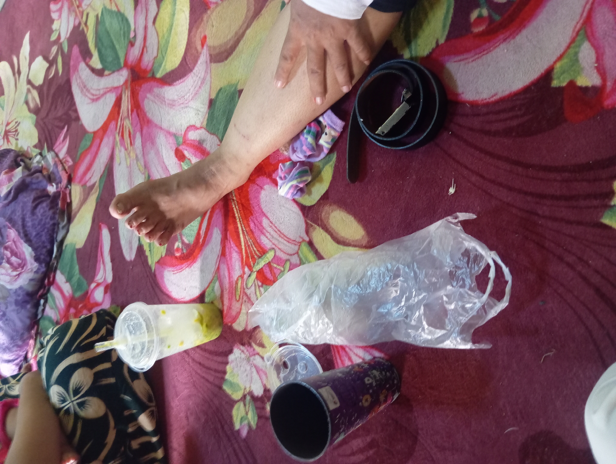 The image depicts a close-up view of a person's leg and foot resting on a fabric with a floral pattern. Several objects are scattered around the leg on the fabric, including a plastic cup with a straw containing a green liquid, a black belt coiled up, two small patterned socks, and a transparent plastic bag. A dark-colored tumbler with a floral design is also visible. The background shows more of the patterned fabric, suggesting an indoor setting possibly on a bed or sofa. The lighting is diffused, and the focus is on the lower body and the objects. The scene does not provide any specific geographical markers to pinpoint a location within Rejosari, Indonesia, but it appears to be a casual, domestic environment.