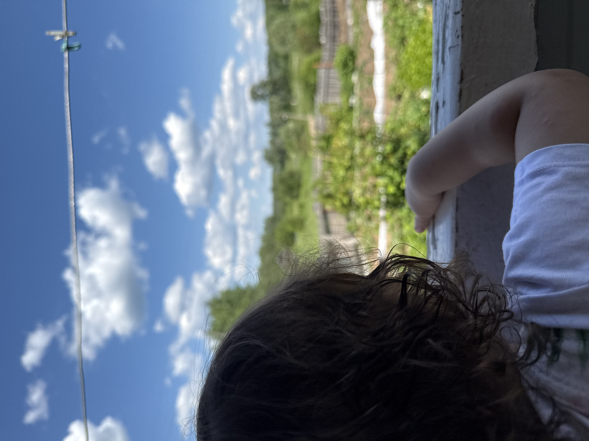 The image shows a child's back and side of their head, looking out over a rural landscape under a bright blue sky with scattered white clouds. The child has dark, curly hair and is wearing a white t-shirt. They are leaning on a weathered, off-white railing, with their right arm bent and resting on it. In the background, there is a field with patches of what appear to be cultivated land and a wooden fence. Trees and greenery are visible in the distance. A clothesline stretches across the top of the frame with a clothespin attached. The scene appears to be outdoors in a village or countryside setting, possibly in or near Уральское, Russia, given the context.