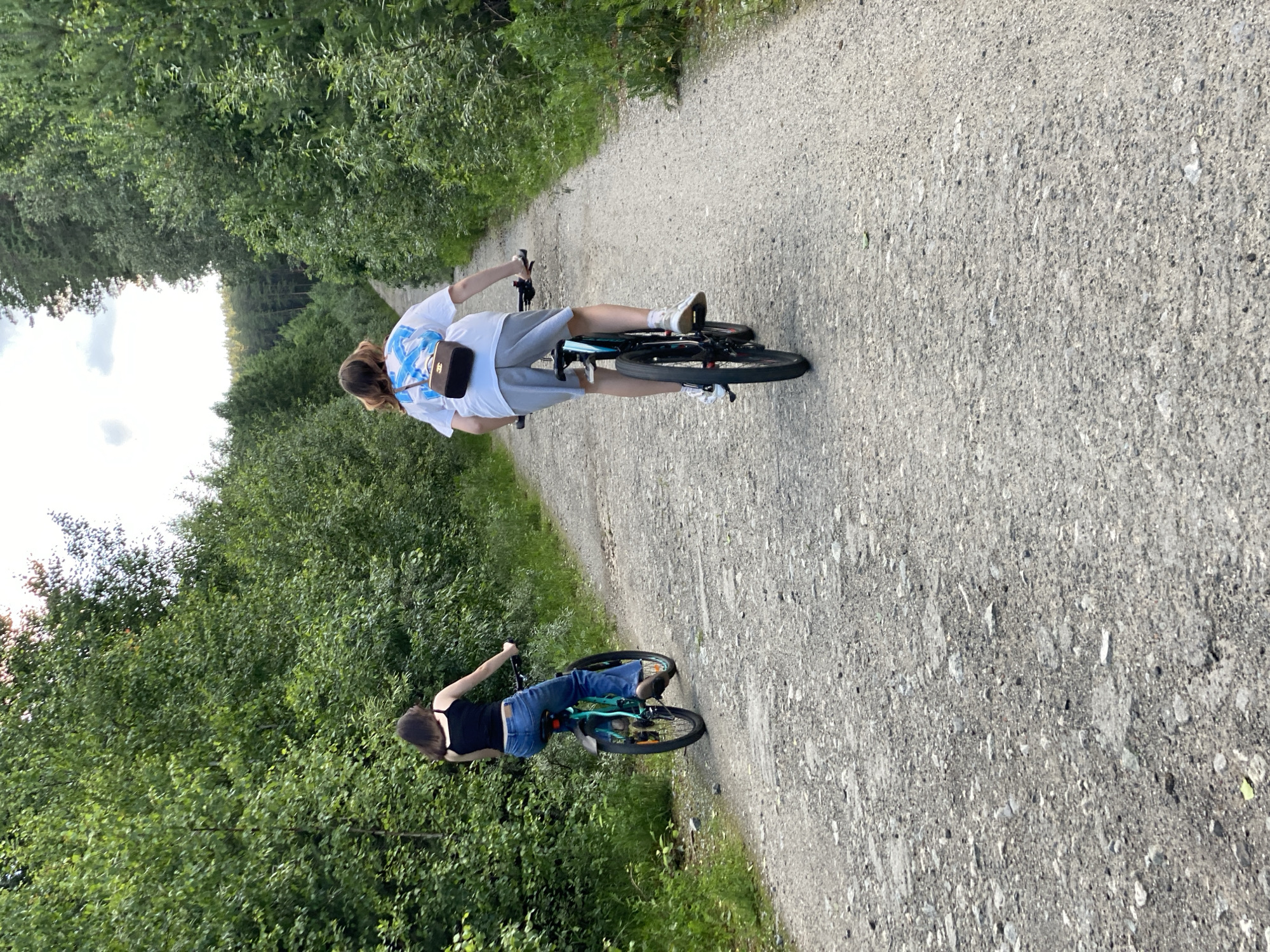 Two individuals are cycling on a gravel path through a forest. The person in front is wearing a white t-shirt with blue accents and grey shorts. They have a small brown bag slung across their chest. The second person, riding behind, is wearing a black tank top and blue jeans.
The scene appears to be a daytime activity, with the lighting suggesting it could be late afternoon or early evening, given the direction of the light and the presence of shadows. The weather seems pleasant for outdoor activity, likely warm and dry. The path is surrounded by dense green foliage, indicating a natural, wooded environment. The overall mood is relaxed and recreational. The image is a still photograph, capturing a moment of leisure. There is no discernible text visible in the image.