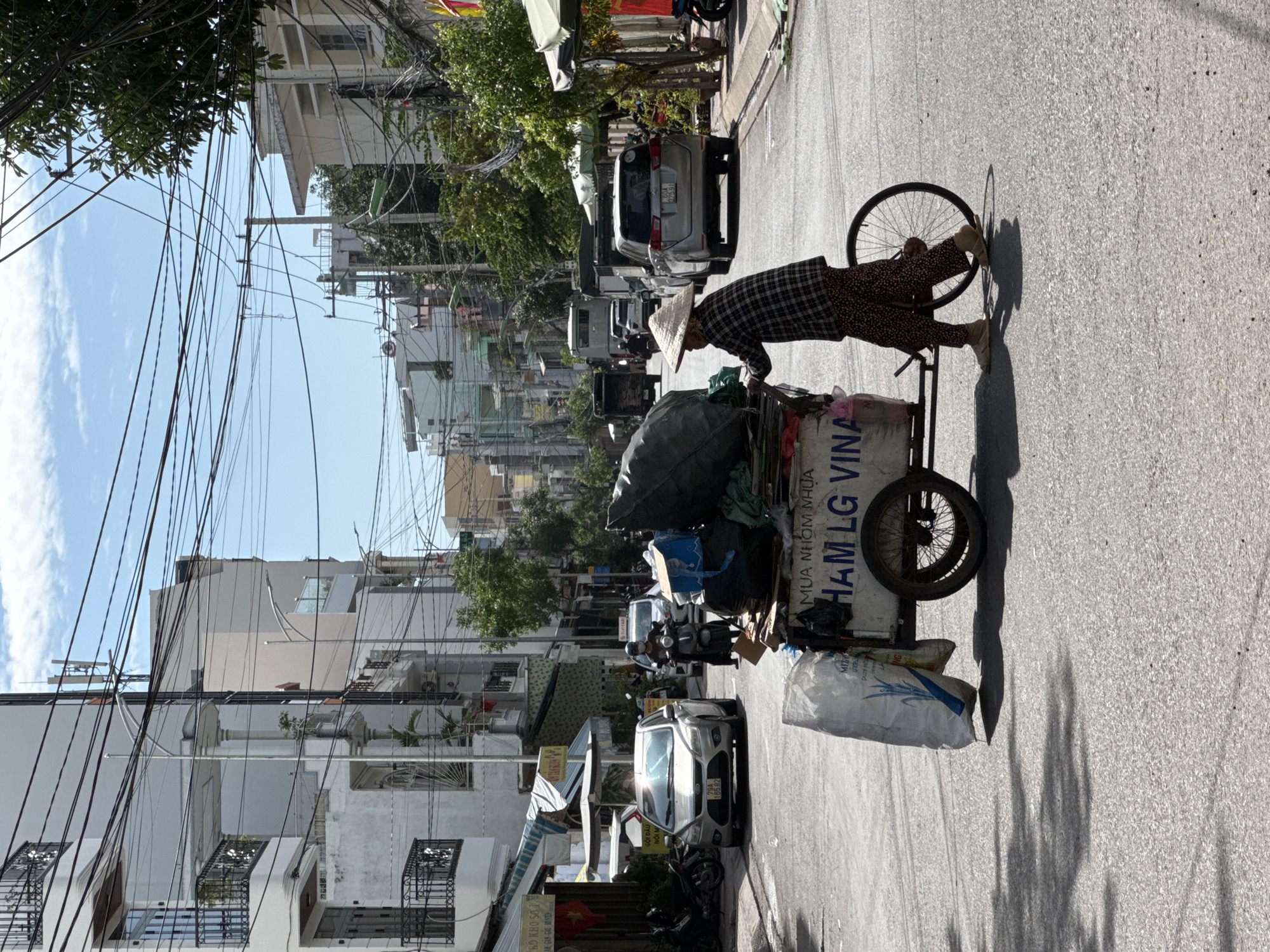 In Nha Trang, Vietnam, a woman wearing a conical hat and a plaid shirt pushes a cart filled with bags and bundles down a city street. The cart has two wheels and appears to be laden with collected refuse or recyclables, indicated by the large plastic bags and the text "MUA NHOM NHUA HAM LG VINA" on its side, which translates to "Buy aluminum plastic HAM LG VINA." She is walking away from the viewer towards the right side of the frame, and her feet are clad in light-colored sandals.
The street is paved and appears to be a typical urban thoroughfare with buildings lining both sides. The buildings are a mix of multi-story residential and commercial structures, some with balconies and intricate architectural details. Overhead, a complex network of electrical wires crisscrosses the sky, a common sight in Vietnamese cities. Sunlight casts distinct shadows of the wires and trees onto the asphalt.
Several vehicles are visible in the background, including a silver car parked on the left, and a scooter with a rider, as well as other cars and trucks further down the street. The overall atmosphere is one of everyday urban life, with the woman and her cart being a central element of the scene. The sky is blue with scattered clouds.