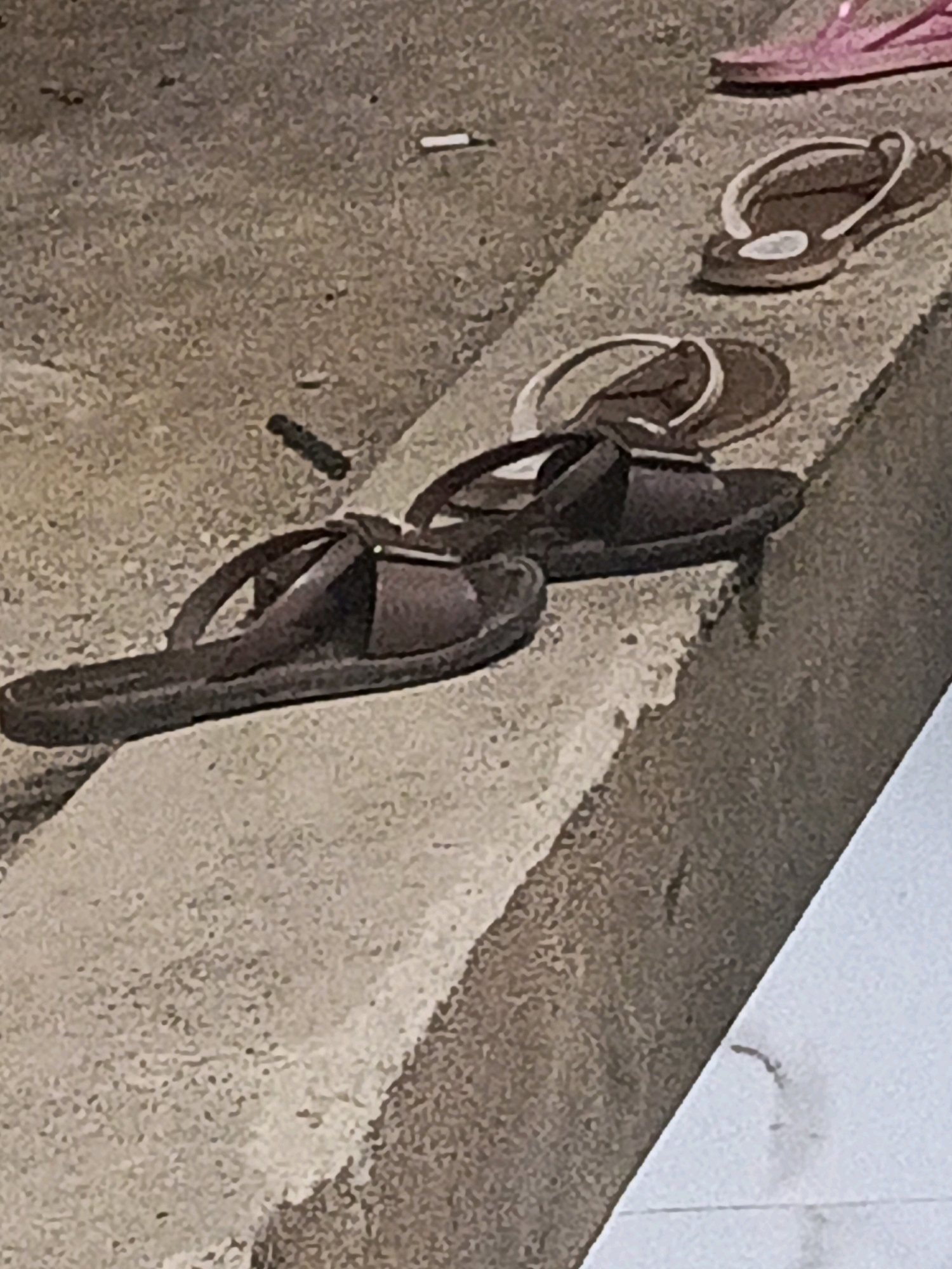 Three pairs of flip-flops are arranged on a textured, grey concrete surface. The closest pair, in the foreground, is dark brown with a thick sole and two straps crossing over the foot. Behind it, further up the incline, are two more pairs of flip-flops. One is lighter brown with white trim, and another, partially obscured by the middle pair, is pink. The concrete surface appears to be a raised walkway or ledge. Small dark debris, possibly cigarette butts or small stones, are scattered on the surface. The overall lighting suggests an outdoor setting, possibly during daylight hours. The context of Megang Sakti V, Indonesia, is not visually represented in the image itself.