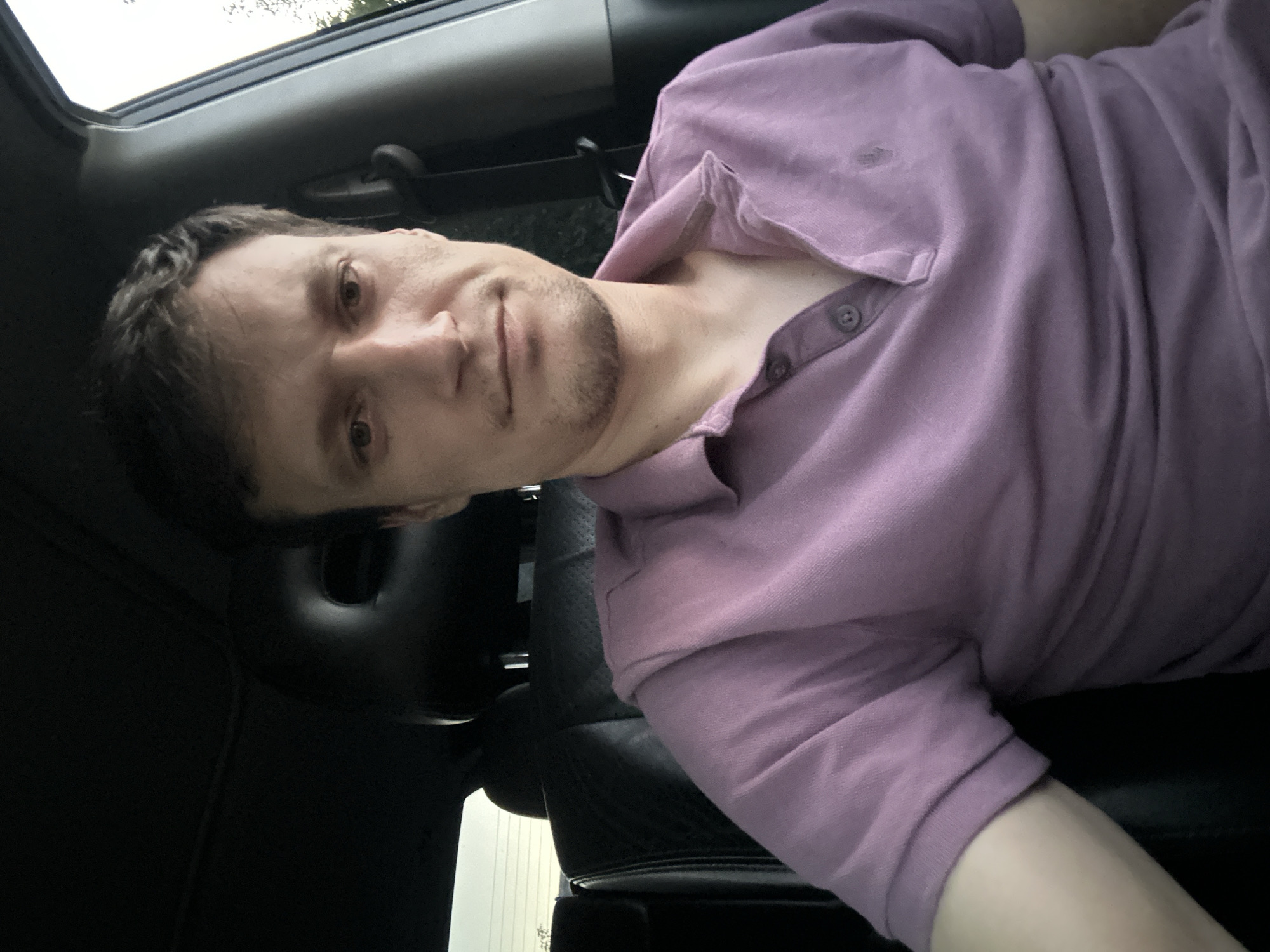A medium shot captures a fair-skinned man with light brown hair and a subtle beard, sitting in the driver's seat of a car. He is wearing a short-sleeved, purple polo shirt. The car's interior is dark, with visible black leather seats and headrests. The background shows the exterior of the car, with a window revealing a hint of the outside environment. This scene is not located in Shatava, Ukraine.