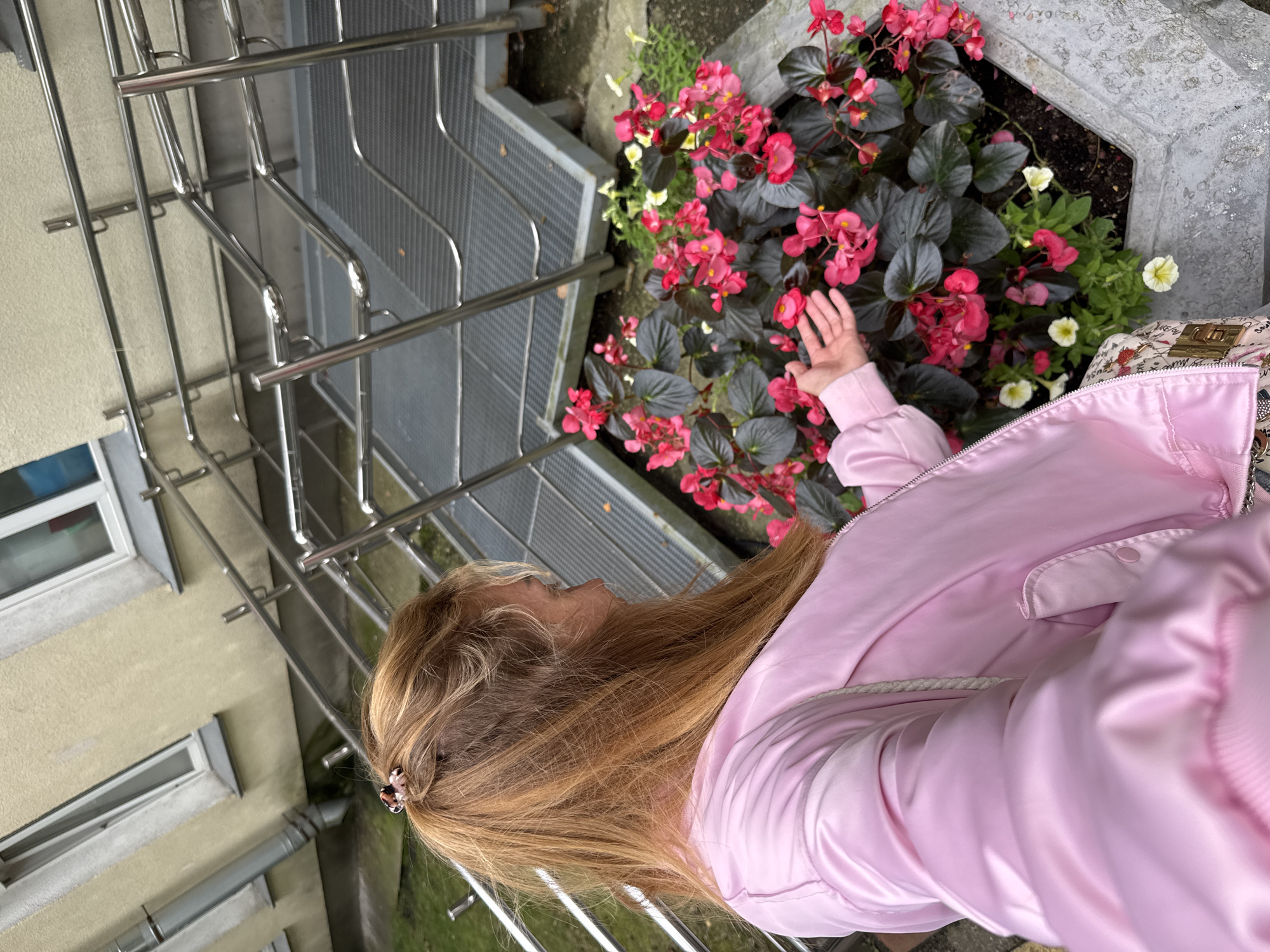 In the city of Sinyavino, Russia, a young woman with blonde hair pulled back by a hair clip stands in front of a planter containing vibrant pink begonias and smaller white flowers. She is wearing a pink satin bomber jacket and a patterned skirt. Her left hand is outstretched, gently touching the begonia flowers. Behind her, a metal staircase with a textured walkway ascends along a building with pale yellow siding and white-trimmed windows. The environment appears to be outdoors, possibly in a residential or public area.