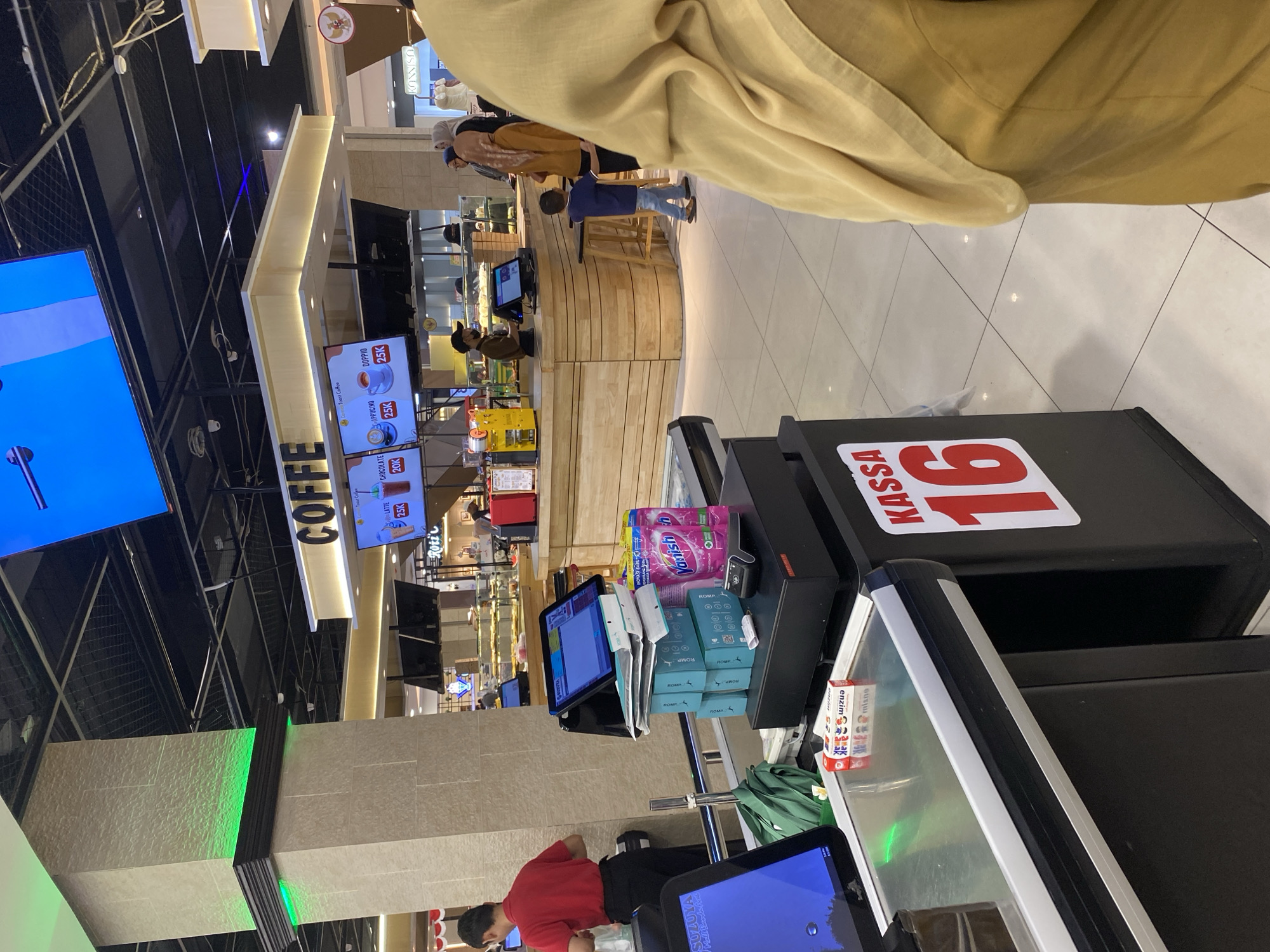 The image depicts the interior of a retail establishment, likely a supermarket or convenience store, in Banda Aceh, Indonesia. In the foreground, a cashier's station is visible, complete with a black cash register, a touchscreen monitor, and various items stacked on it, including boxes and a pink detergent packet. A large red and white sign with the text "KASSA 16" is prominently displayed on the side of the cash register. To the left, another monitor is partially visible, showing a blue screen.
In the middle ground, several individuals are present. A person wearing a red shirt and black pants, possibly a store employee, is seen from the back, facing away from the camera and interacting with items behind a counter. A young child in a blue shirt and jeans is standing near a wooden counter area, which displays baked goods or other food items. Other shoppers, including two women wearing headscarves, are also in this area.
In the background, a "COFFEE" sign with illuminated text is visible above a counter area that appears to be a coffee or food service station. Digital menu boards are also displayed, showing drinks and prices. The overall environment is well-lit, with a modern ceiling structure and tiled flooring. The presence of the Indonesian flag on a circular emblem in the background further suggests the location is within Indonesia.