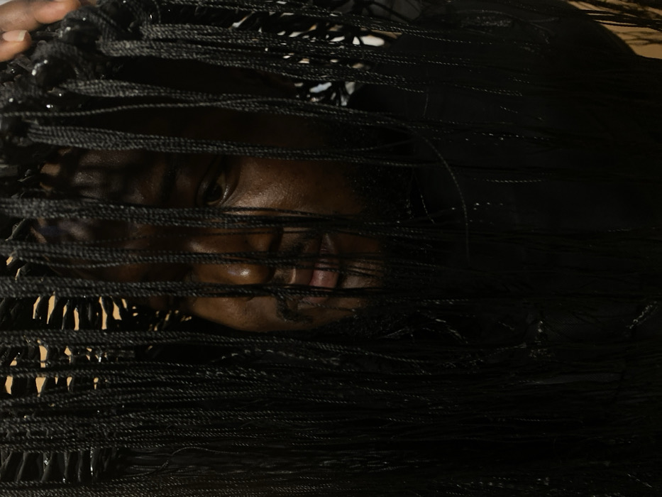 A medium-close-up portrait features an individual with dark skin, exhibiting male characteristics including a beard and mustache. The person's face is partially obscured by numerous thin, vertically hanging black braids, appearing to be either a hairstyle or a decorative element. The right eye (viewer's left) is visible and directed slightly downwards, with a neutral expression. A dark-skinned hand is present at the upper right, appearing to adjust or support the braided material. The individual wears a dark-colored garment. The background is dark and indistinct, with faint glimpses of a lighter, possibly textured surface visible through the gaps in the braids at the top. No specific location within Jos, Nigeria, is identifiable from the image.