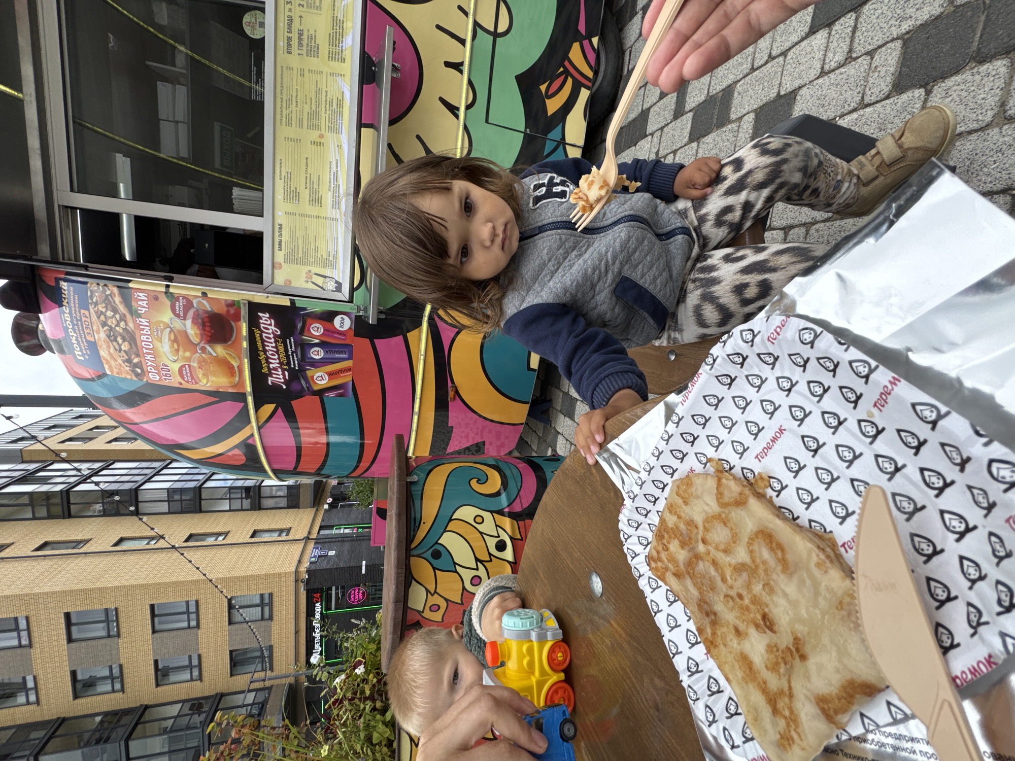 A young child is seated at a wooden table outside a food truck, with a portion of a blini on a paper wrapper in front of them. The child is looking at a fork held by an unseen person, which has food on it. To the left, another child's hands are visible, playing with a toy train. The food truck has a colorful, abstract design and displays a menu board. In the background, a modern building with a yellow brick facade and large windows is visible. The setting appears to be an outdoor urban area, possibly a food court or street-side dining area, in Novoselye, Russia.