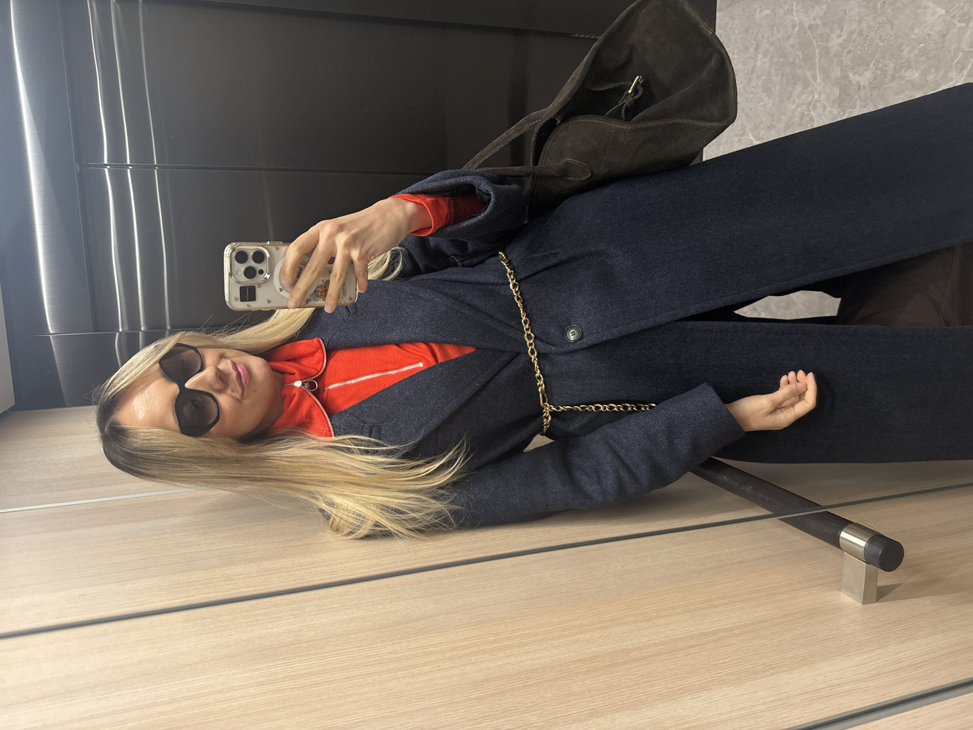The image, taken in Riga, Latvia, depicts a woman posing for a mirror selfie in an indoor setting. The image is rotated to a horizontal orientation, with the woman lying across the frame. She has long, straight blonde hair and is wearing dark, oversized sunglasses, which obscure her eyes.
Her attire includes a long, dark navy blue coat, possibly wool or a similar textured fabric, worn over a bright orange long-sleeved top with a visible quarter-zip neckline. A thin gold chain belt is draped around the waist of her coat. She is holding a light-colored smartphone, likely an iPhone, in a clear case adorned with several small decorative items and a circular attachment on the back. A dark brown, possibly suede, shoulder bag is visible slung over her shoulder behind her.
The background consists of a reflective dark surface, resembling polished metal or a mirror, on the upper side of the frame, and a light-colored wooden floor on the lower side. A dark cylindrical object with a silver band and cap, possibly a cane or a part of interior decor, rests on a small, light-colored rectangular base on the floor. The overall setting suggests a modern interior space, such as a hallway or an elevator. The woman's pose and fashionable attire indicate a casual outfit-of-the-day or style photo.