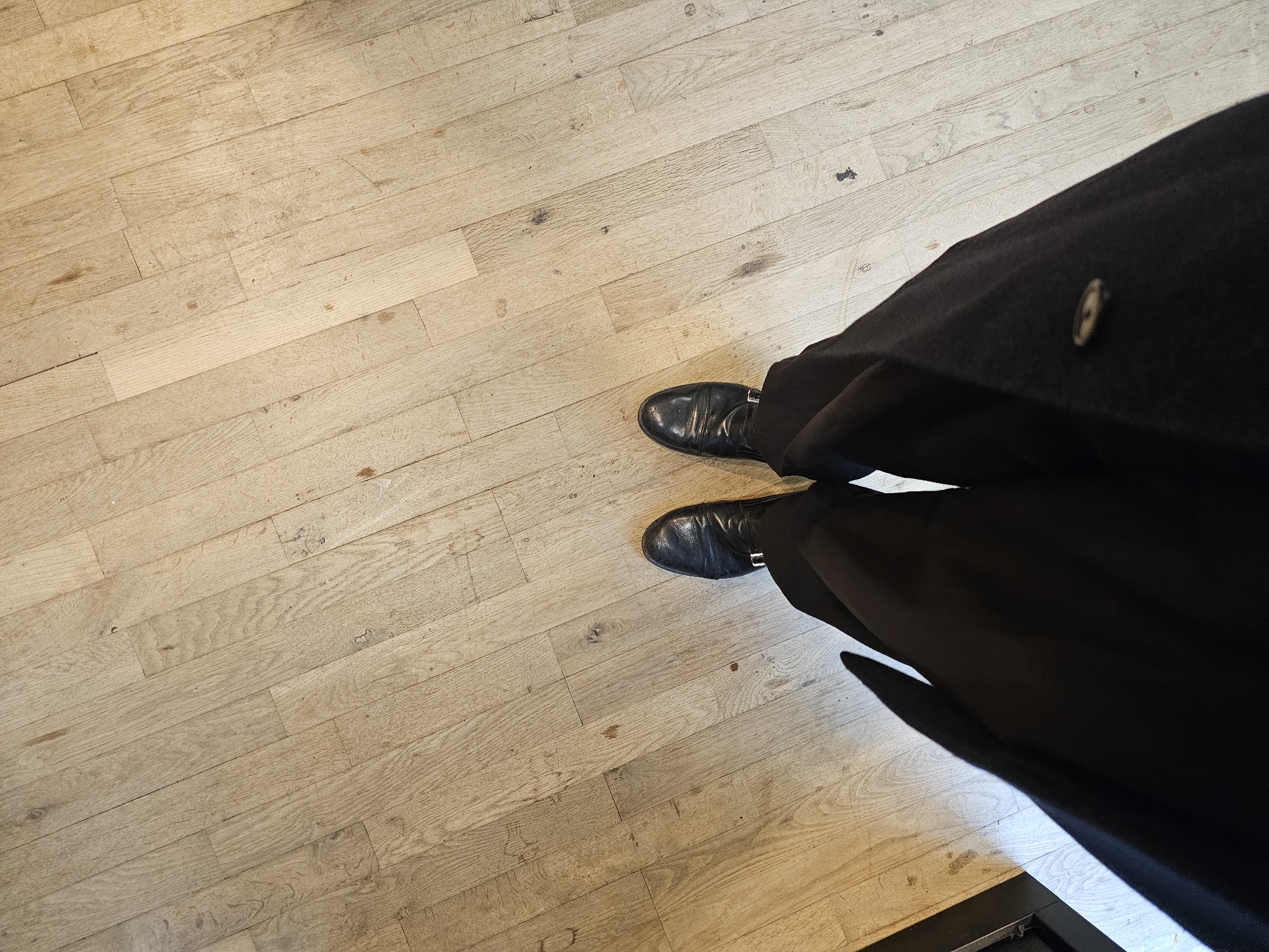 A first-person perspective photograph depicts a person standing on a light-colored, wooden plank floor. The person is wearing black dress shoes and black trousers with a long black coat or jacket extending down towards the right side of the image. The floorboards run diagonally from the top left to the bottom right. A partial view of what appears to be a metal frame can be seen in the bottom right corner. The location is likely indoors, but there are no other identifying environmental details visible in the photograph to place it specifically in Paris.