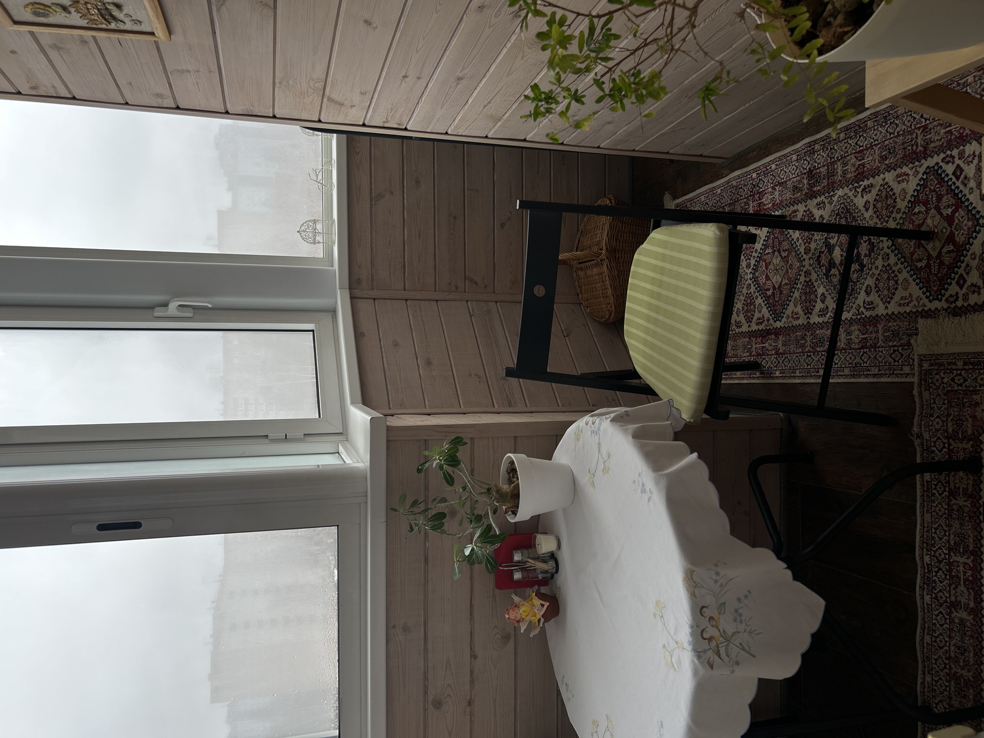 The image depicts a small, enclosed balcony or loggia. The walls and ceiling are clad in light-colored, horizontal wooden planks. A white-framed, multi-pane window is on the left, showing a view of a cityscape obscured by overcast weather. The buildings appear to be typical of an urban environment.
A small, round table covered with a white tablecloth with floral embroidery is in the lower portion of the image. A potted plant sits on the table, next to a red container holding kitchen utensils, and a small figurine.
A black chair with a striped green cushion is positioned near the table. The chair's back is against the wooden wall.
Part of a patterned rug is on the floor. A wicker basket is visible between the chair and the wall.
Based on the architecture visible through the window and contextual evidence, it could be hypothesized that the location is Izhevsk, Russia, but without further corroborating information, this identification remains speculative. The scene is static and contains no people.