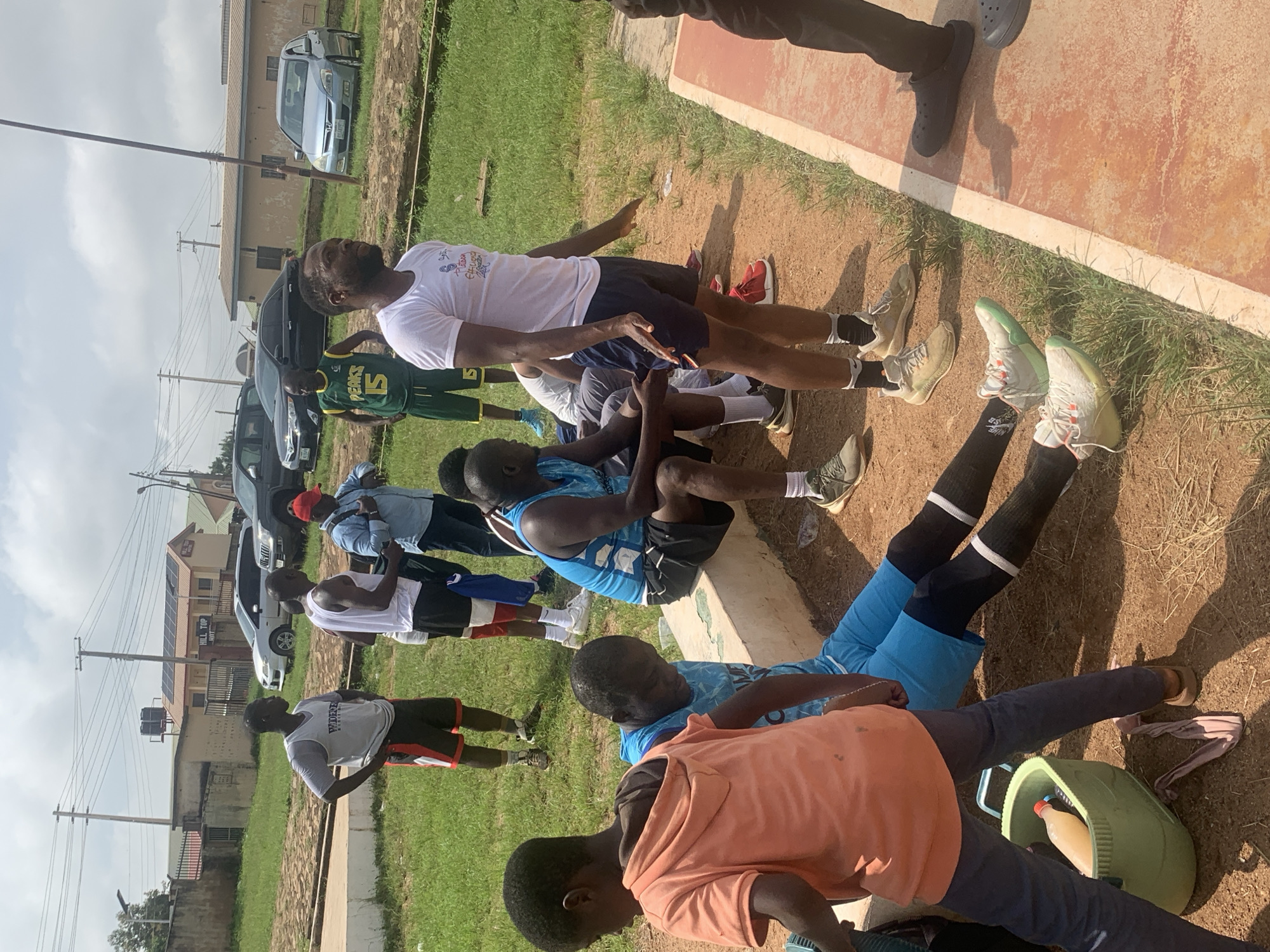 In Jos, Nigeria, under a partially cloudy sky, a group of men and boys are gathered outdoors on a dirt and grassy area. Some individuals are standing, including a man wearing a white t-shirt and dark shorts, another in a grey tank top and red and black shorts, and a man in a green and yellow tank top with the number 15 on it. Other individuals are seated, including a man in a blue tank top and black shorts, and a young boy wearing an orange hoodie. A larger young boy in an orange hoodie is visible in the foreground. Several cars are parked in the background, including a silver sedan, a black SUV, and a light blue sedan. A building with solar panels on its roof is also present in the background. An assortment of athletic wear, including shorts and tank tops, is being worn by the people in the image. A green tub containing a bottle of liquid is on the ground near the foreground.