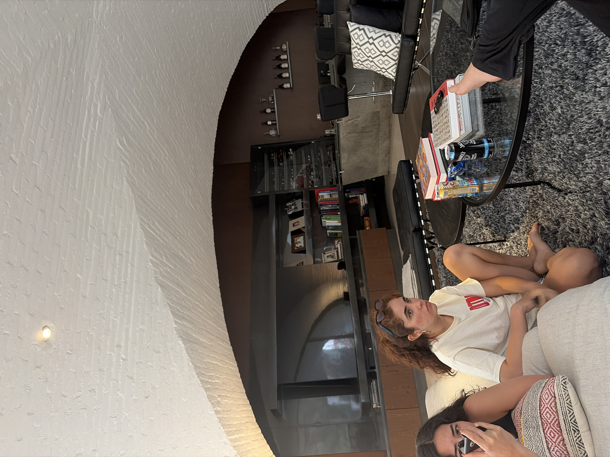 A slightly high-angle interior shot shows two young women lounging on a couch. One is lying on her side, wearing a white T-shirt, and gazing toward the upper left corner of the frame. The other woman is sitting more upright and looking down while holding a smartphone. A patterned pillow sits behind her. A glass-topped table, cluttered with books and cans, sits in the upper right of the frame. Behind the women and the table, a series of shelves is visible, populated with books and decorative objects. In the background, there's a unique, arched architectural design element, featuring a white brick wall that curves into the upper part of the frame. Some overhead lighting fixtures are also noticeable on the wall. The scene is indoors, possibly in a living room setting.
