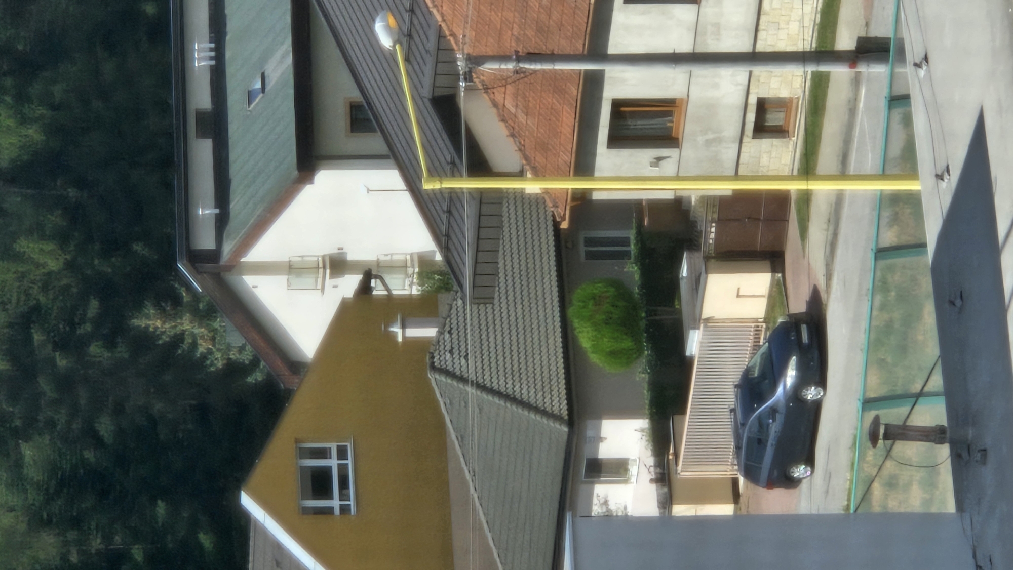 A low-angle, eye-level shot captures a street scene in Prešov, Slovakia. In the foreground, a dark blue minivan is parked on a paved road. The minivan has a roof rack and its headlights are on. Behind the minivan, a light-colored fence surrounds a property with a small, rounded green shrub and a gate. Several buildings are visible in the background. To the left, a yellow house has a large, white-framed window. Adjacent to it is a white building with a green roof. Further to the right, a grey building with an orange-tiled roof is partially visible, featuring a prominent yellow lamppost. The environment appears to be residential, with dense trees forming a backdrop to the buildings. The lighting suggests it is daytime.