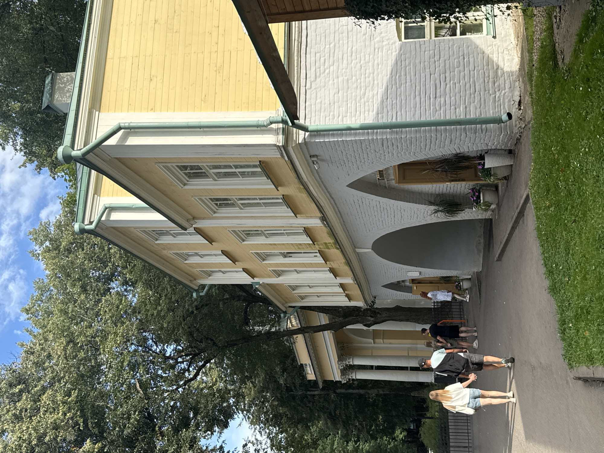 The image depicts the historic museum estate of Arkhangelskoye, located in the village of posyolok dacha khozyaystva "Arkhangelskoye", Russia. The central subject is a two-story building with a striking yellow upper facade and a white brick lower section featuring arched openings. The architecture exhibits classical influences with white trim around windows and pilasters on the adjacent colonnaded structure. Lush green trees frame the scene, suggesting a park-like setting.
In the foreground, a couple is walking away from the camera, holding hands. The man is wearing a black cap, a light blue t-shirt, and dark shorts. The woman is in a white t-shirt and light denim shorts. Behind them, a group of four people are also walking, their details less distinct. Further to the right, a woman in a white dress is visible near the arched entrance of the building. Several potted plants are placed near the archways, adding a touch of greenery. The sky above is partly cloudy, with patches of blue. The overall impression is a sunny day in a historical and serene location.