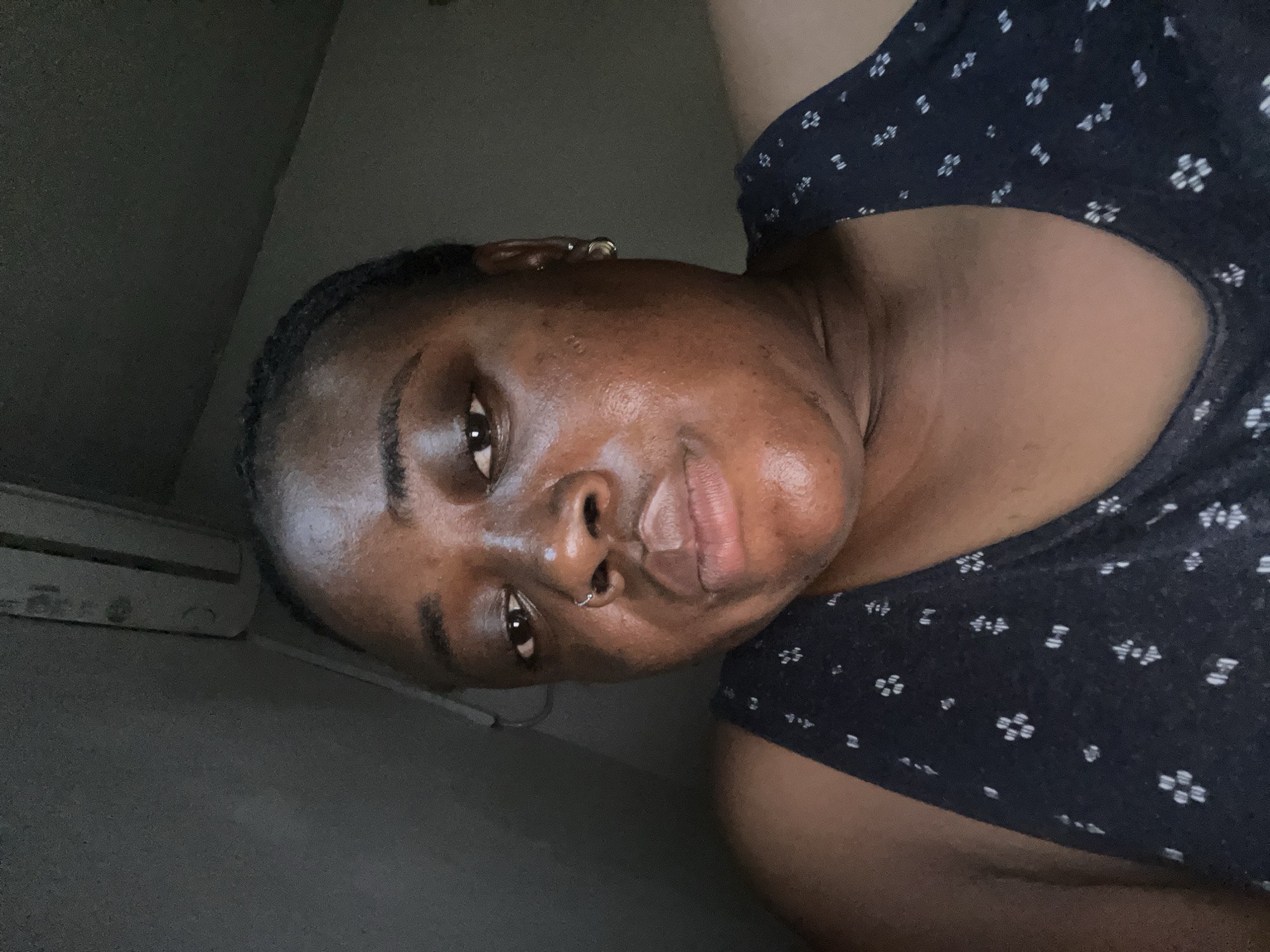 A medium close-up shot of a dark-skinned Black woman wearing a dark blue shirt with a white floral pattern. She has short dark hair pulled back from her face, and a nose ring. The background appears to be a dimly lit room, possibly in a residence, with a wall and an air conditioning unit visible. It is not possible to determine the location within Abuja, Nigeria from the image.