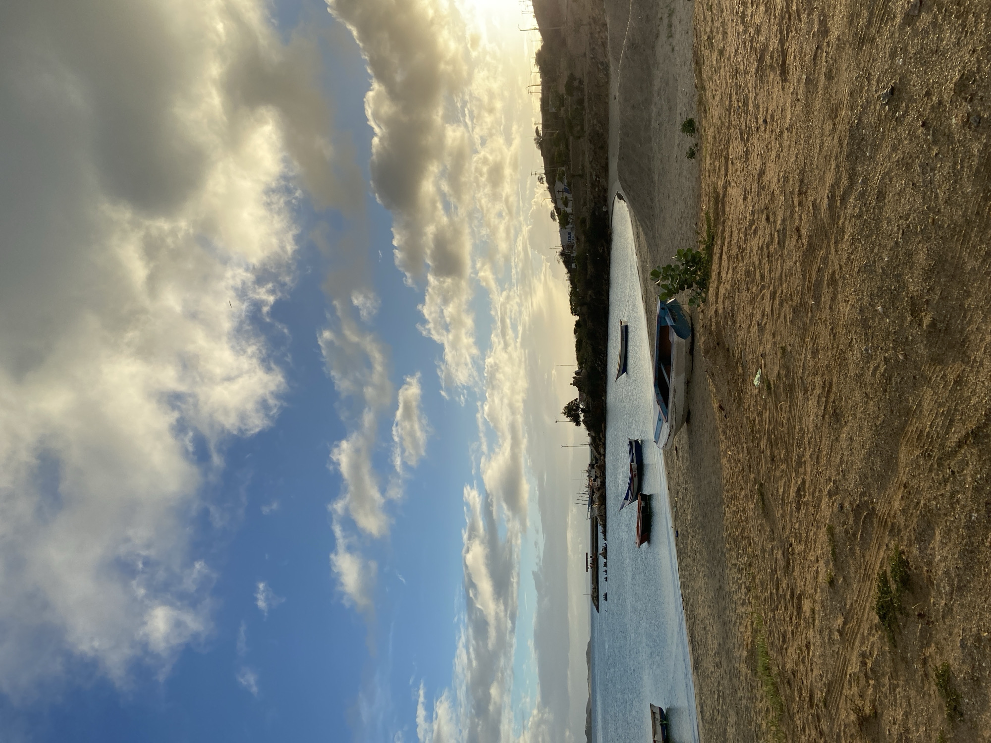 This is a photograph of a beach in Juan Griego, Venezuela. The scene is dominated by a sandy beach in the foreground, with tire tracks visible in the sand. Several small boats are pulled up onto the beach, and one is partially submerged in the shallow water near the shore. The water stretches out to the horizon, meeting a sky filled with dramatic clouds. In the distance, a rocky shoreline with sparse vegetation and a few buildings can be seen. The sun is low in the sky, casting a warm glow and creating long shadows. There are no people visible in the image.