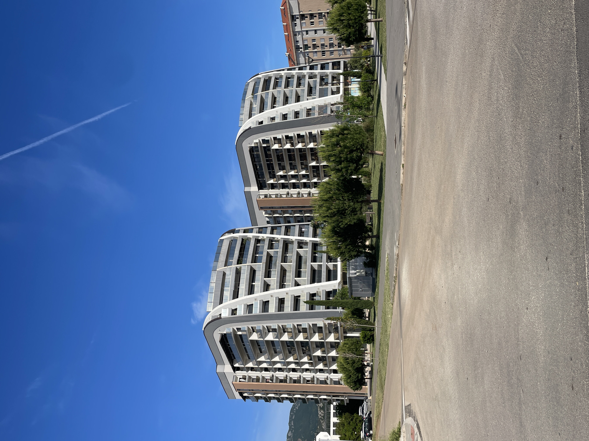 The image shows two modern, high-rise apartment buildings in Bar, Montenegro. The buildings are predominantly white with grey accents, featuring a curved design. A clear blue sky with a contrail overhead serves as the backdrop. A wide asphalt road is in the foreground. Some trees and greenery line the sidewalk between the buildings and the road. A mountain or hill is visible in the far distance, behind the apartment buildings.