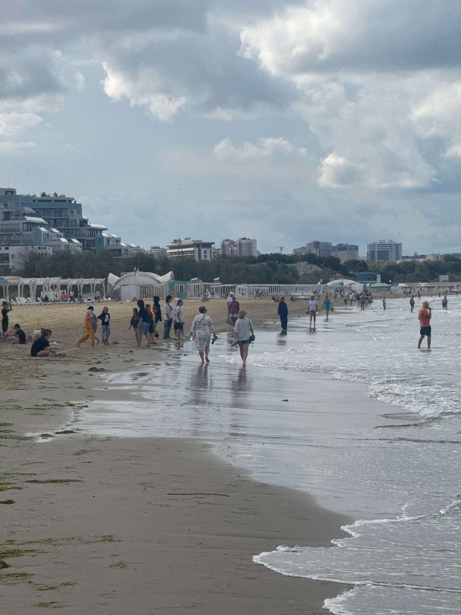 A group of people are gathered on a sandy beach with the sea lapping at the shore. Buildings line the background, and a cloudy sky looms overhead. The scene appears to be the beach in Anapa, Russia, a popular resort town.