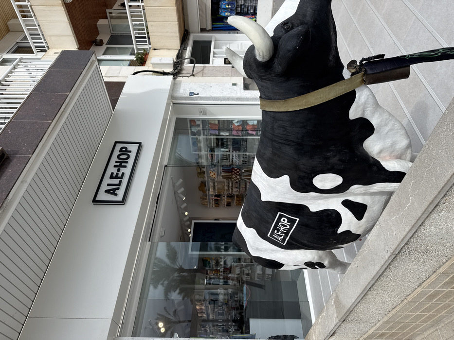The image depicts a black and white Holstein cow statue positioned on the sidewalk in front of an "ALE-HOP" store in Port D'alcúdia, Spain. The statue, facing right and partially obscured by a low stone wall in the foreground, features the "ALE-HOP" logo in a white rectangular box on its left flank. It wears a tan-colored strap around its neck, from which a dark bell-like object and a green-patterned strap are suspended.
The "ALE-HOP" store entrance features a white facade with a black-framed, illuminated sign displaying "ALE-HOP" in white letters centrally above large glass automatic sliding doors. Through the glass, the brightly lit store interior is visible, showcasing various merchandise displays, including shelves stocked with items and what appears to be a rack of wigs or hair accessories on the left. Overhead track lighting illuminates the interior. Reflections in the glass doors include palm trees and ambient outdoor lighting.
Above the store, a multi-story building with balconies is visible. The upper facade consists of dark brown textured panels and white railings, with some wooden accents on the lower balcony of the building above the store. A dark cable runs vertically down the white facade next to the store entrance. The sidewalk in front of the store is paved with light-colored tiles, and a gray stone curb or planter separates the immediate foreground from the main walkway. No human figures are present in the immediate foreground or directly interacting with the cow statue or store entrance.
