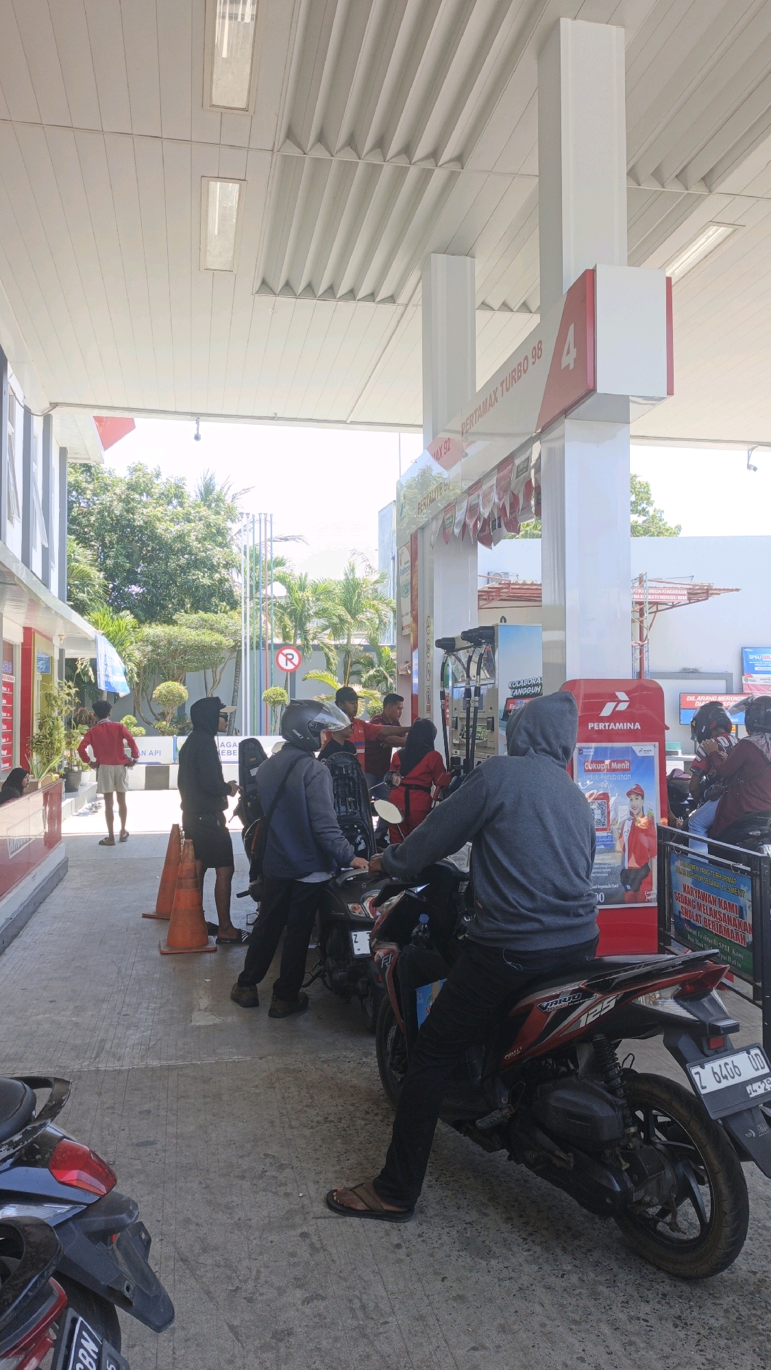 The image depicts a busy scene at a Pertamina gas station, likely located in Pangandaran, Indonesia. Several individuals on motorcycles are queuing to refuel. The station has a white, modern canopy supported by pillars. Prominent signage indicates "PERTAMAX TURBO 98" and other fuel types. The ground is paved concrete, with orange traffic cones placed near a queue of motorcycles. People are dressed in casual attire, with some wearing hoodies and helmets. Two individuals in red uniforms, likely gas station attendants, are visible near the pumps. The environment suggests a warm, sunny day with trees and palm trees visible in the background. A blue sign with text in Indonesian is mounted on the gas pump. The overall atmosphere is one of everyday commerce and activity.