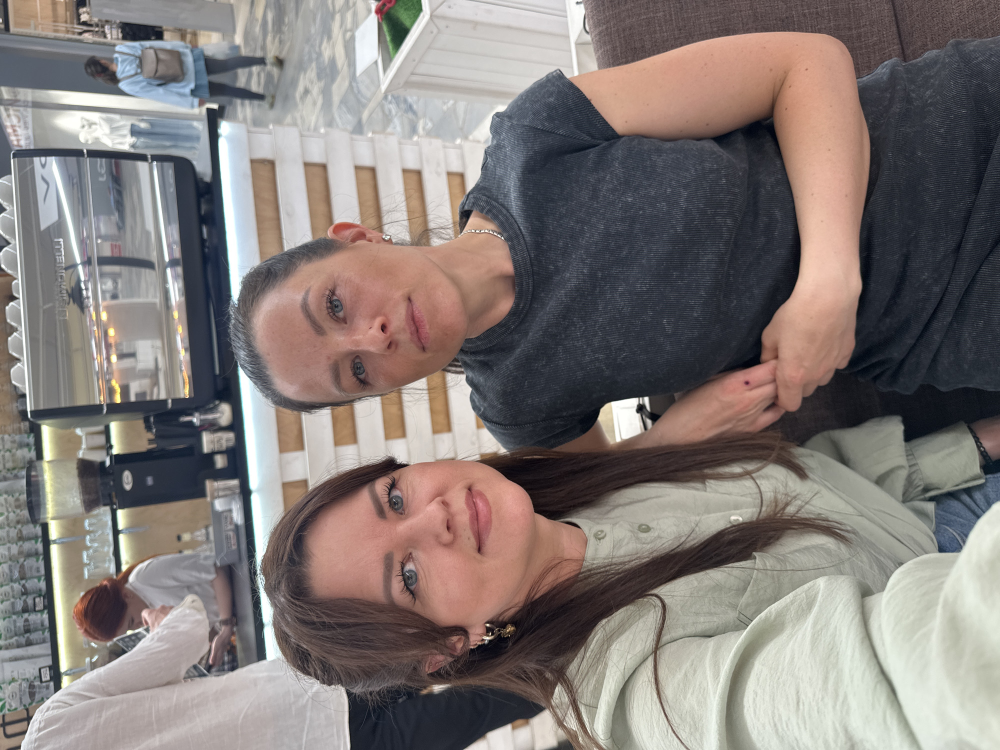 Two women are posing for a selfie indoors. The woman on the left wears a gray t-shirt and a silver necklace. The woman on the right wears a light green shirt and has long brown hair. A coffee machine and a cafe worker are visible in the background. The location could be a cafe in Chaikovsky, Russia, based on the prompt. The women appear to be sitting together.