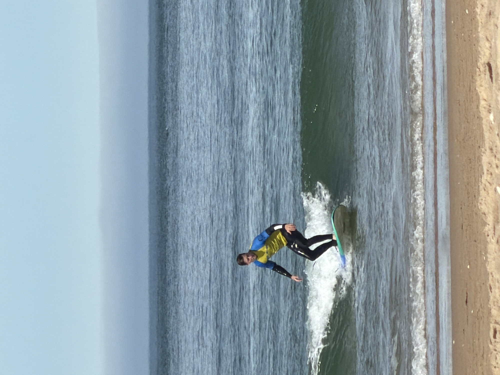 An athletic man, wearing a yellow and blue wetsuit, is surfing a wave near the shore in Almada, Portugal. He is standing on a light blue surfboard and is mid-ride, with a wave cresting behind him. The ocean is a deep blue, and a sandy beach is visible in the foreground. The sky is clear with a hazy layer of fog on the horizon.