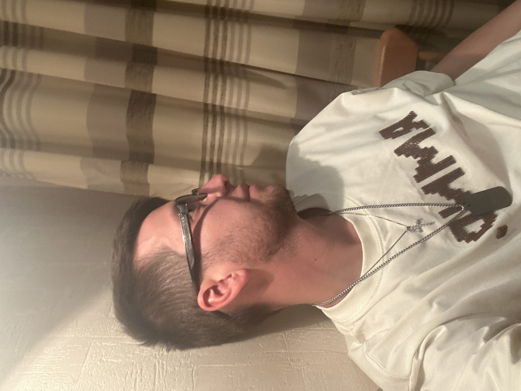 A medium shot captures a young man from the chest up, facing right and leaning against a textured wall. He is wearing a white t-shirt with a dark, pixelated graphic that appears to spell "GAMMA". Two silver necklaces are visible around his neck, one with a cross pendant and the other with a dog tag. The man has short, dark hair, stubble, and is wearing glasses with dark frames. The background consists of striped curtains in shades of beige and brown, and a hint of a wooden chair. The lighting suggests an indoor setting, possibly a home, with a shadow cast on the wall from an unseen light source. This scene is located in the city of Khimki, Russia.
