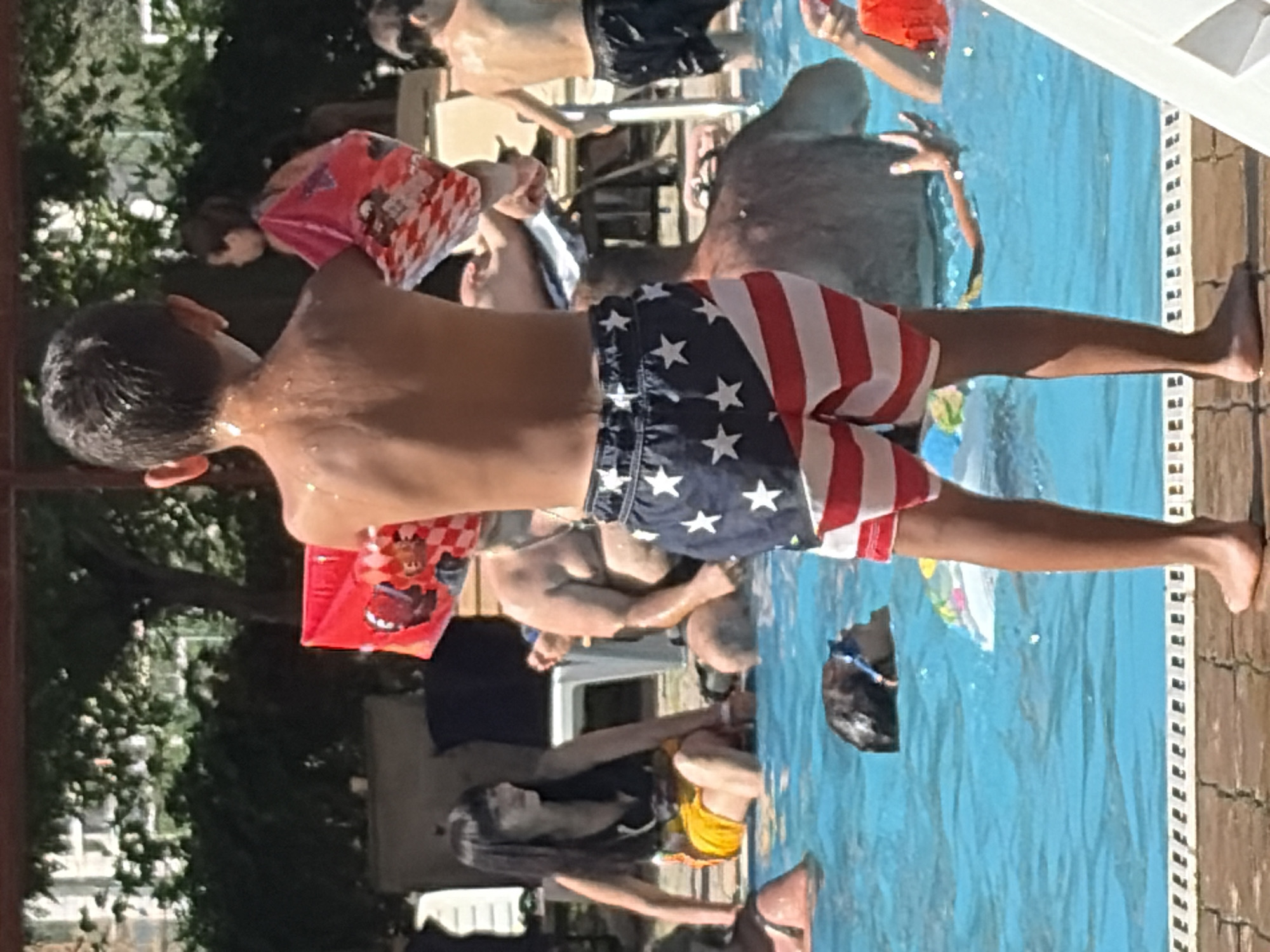 The image shows a group of people at a swimming pool on a sunny day. In the foreground, a young boy stands with his back to the camera, wearing American flag-themed swim trunks. He is holding a red and white checkered toy. To his left, a woman with long dark hair sits in the pool wearing a black tank top and yellow shorts. To the right of the boy, a man with a shaved head and a large build is partially visible in the pool, and further back, another young person in a swim suit is also in the water. Several other individuals are visible in and around the pool, including a child with a dark head of hair submerged in the water. The pool has bright blue water, and the surrounding area is paved with brick. Lush green trees and part of a building are visible in the background.