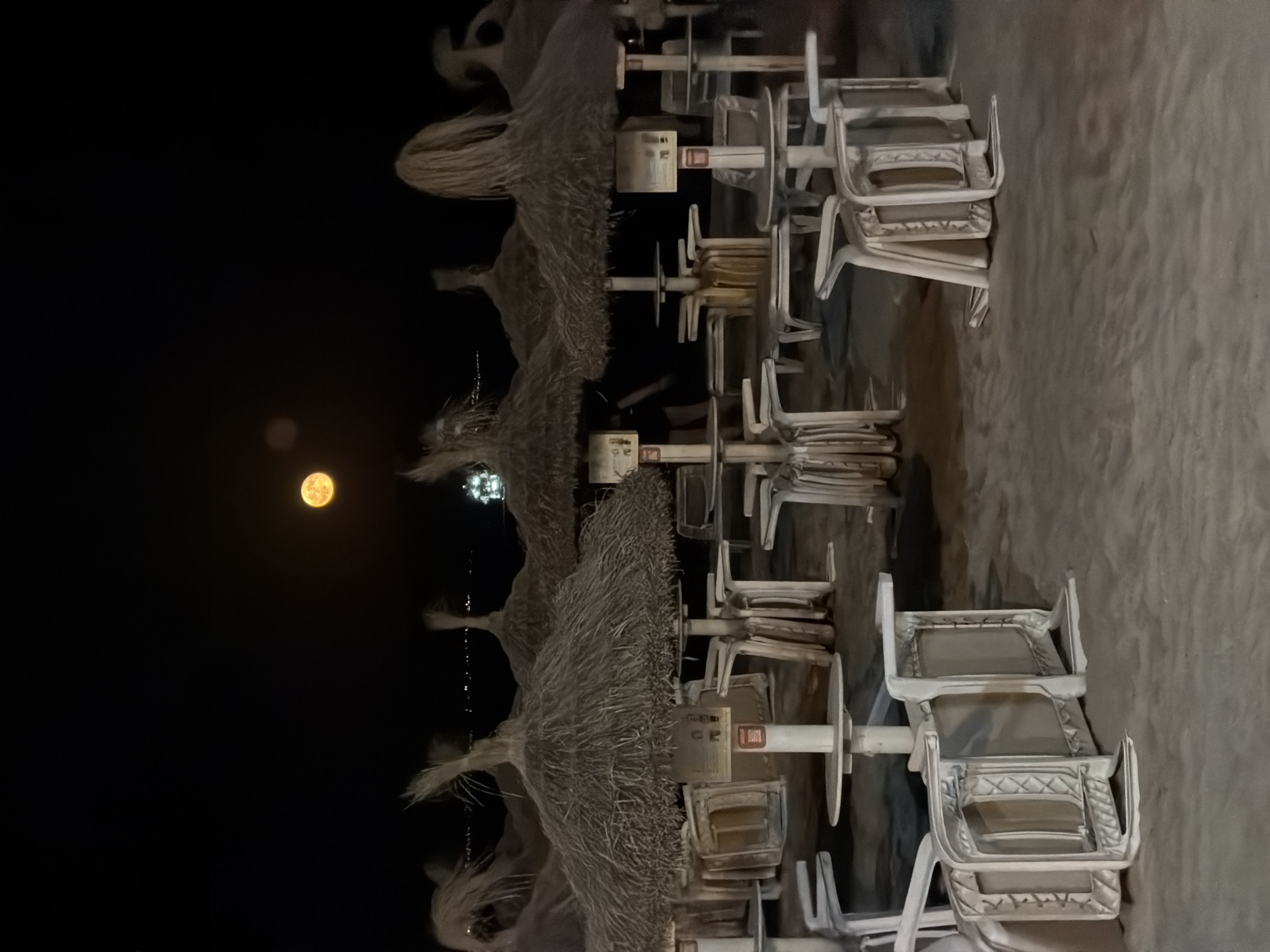 The image shows a beach at night in Palmanova, Spain. The moon is visible in the sky, and there are several beach umbrellas and tables set up on the sand. Many of the chairs are overturned or stacked, suggesting the beach is closed for the night. The overall scene is dark and quiet, with the moon providing the primary illumination.