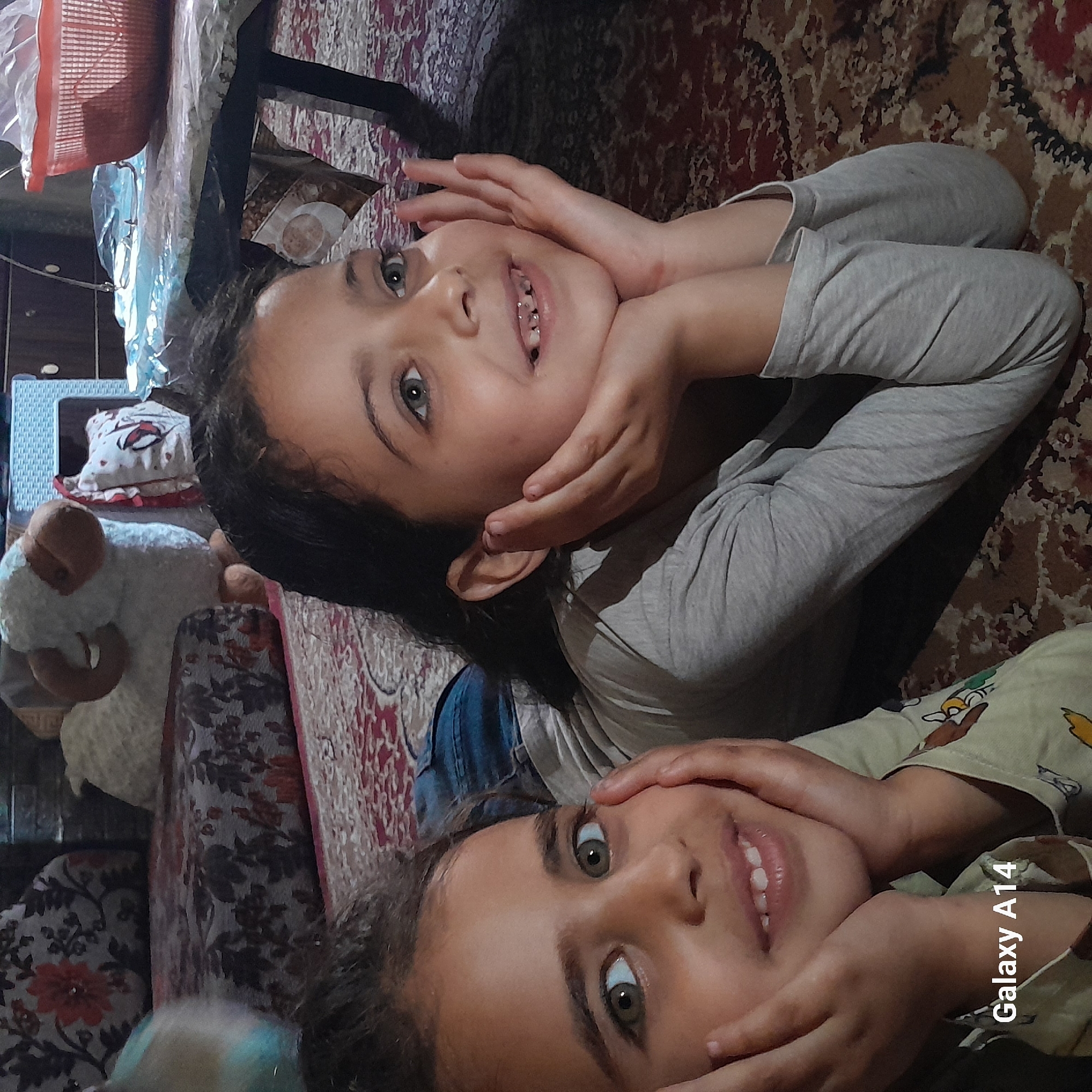 Two young girls are lying on a patterned rug, propped up with their hands under their chins, smiling at the camera. The girl on top is wearing a gray long-sleeved shirt and the girl on the bottom is wearing a light-colored, patterned long-sleeved shirt. The rug has a predominantly brown and red pattern. In the background, there is furniture with items on top, including a basket and a stuffed animal. The image also has the watermark "Galaxy A14". The location is unknown.