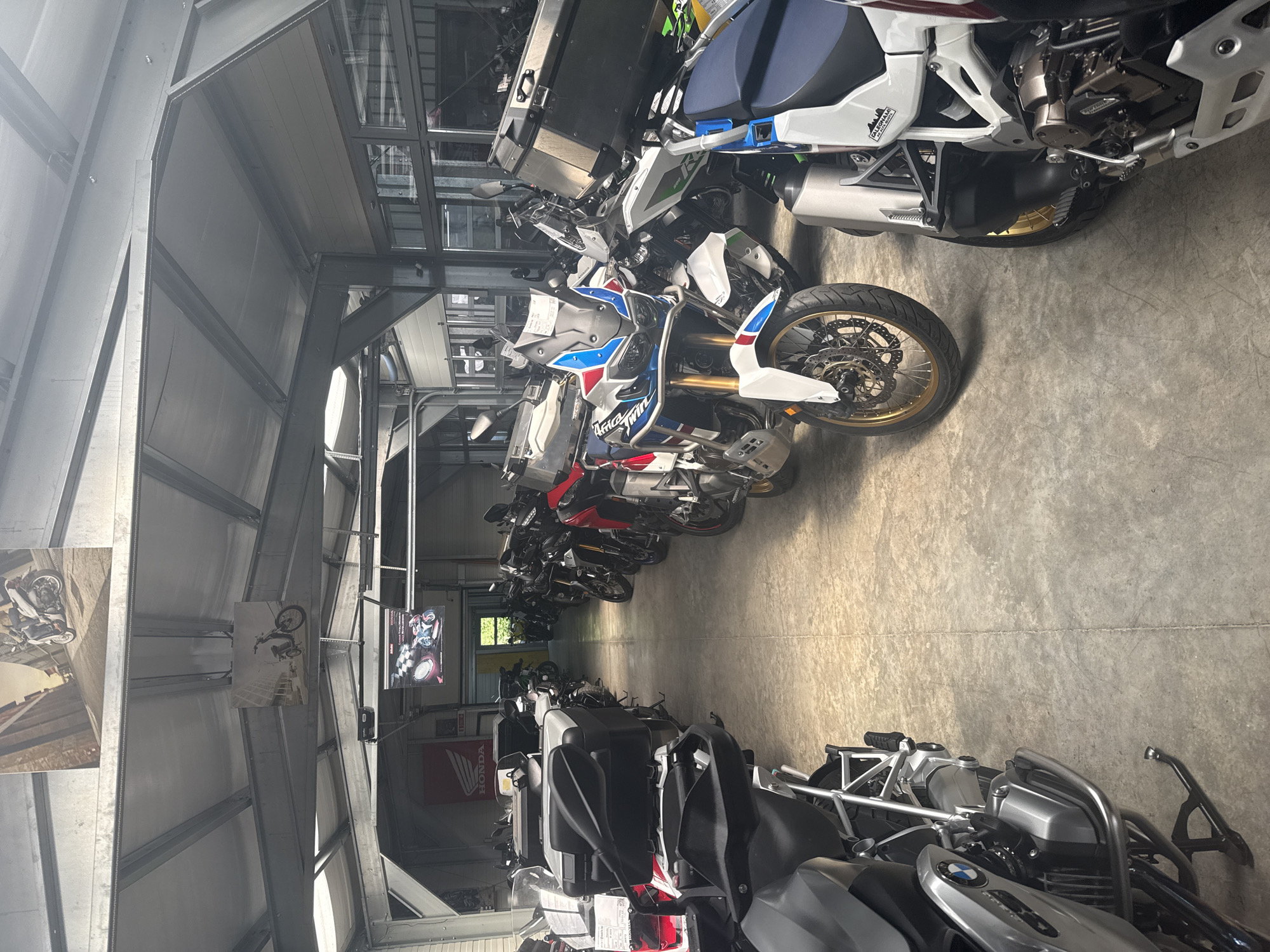 The image displays the interior of a motorcycle showroom or large garage, located in Piperevo, Bulgaria. The space is filled with numerous motorcycles, predominantly adventure-style models, neatly lined up along a concrete floor. No people are visible in the scene.
In the foreground, a prominent white, blue, and red Honda Africa Twin motorcycle is visible, with "Africa Twin" text clearly displayed on its fairing. Just below it, a "KLEEMANN" sticker is also noticeable. Another BMW motorcycle is partially visible in the bottom right, identifiable by its BMW logo on the engine casing. A long row of additional motorcycles, some equipped with top cases, extends into the background, suggesting a substantial inventory or collection.
The building exhibits an industrial aesthetic with a high ceiling featuring exposed metal beams and support structures. Large windows or glass panels are visible at the top, allowing for natural light, complemented by overhead artificial lighting. Motorcycle-themed posters adorn the walls and structural elements; one clearly depicts a trials rider in mid-air, while another partially visible poster suggests a racing theme. A red banner with the "HONDA" logo is also visible in the mid-ground. The concrete floor and robust structural components indicate a functional, commercial, or storage setting for the vehicles.