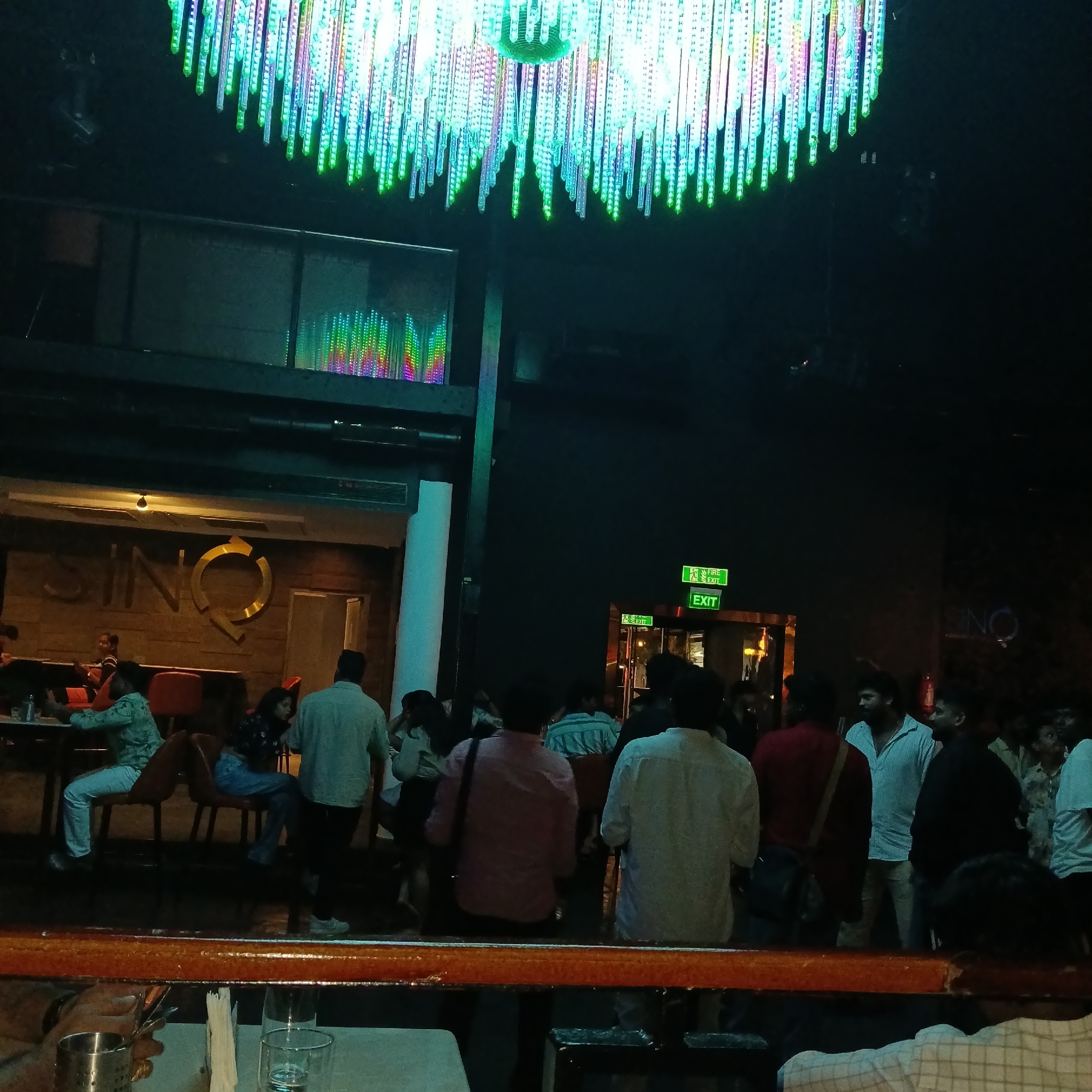 A dimly lit interior shot shows a crowded bar or nightclub, possibly located in Sinquerim, India, suggested by the "SinQ" signage visible on the wall. A large, decorative lighting fixture with multi-colored LED strands hangs from the ceiling. Several people are seen standing and sitting around tables. An "EXIT" sign is visible. There is a bar or counter in the foreground, with glasses and tableware present. People appear to be socializing and conversing. The overall environment suggests a lively nightlife setting.