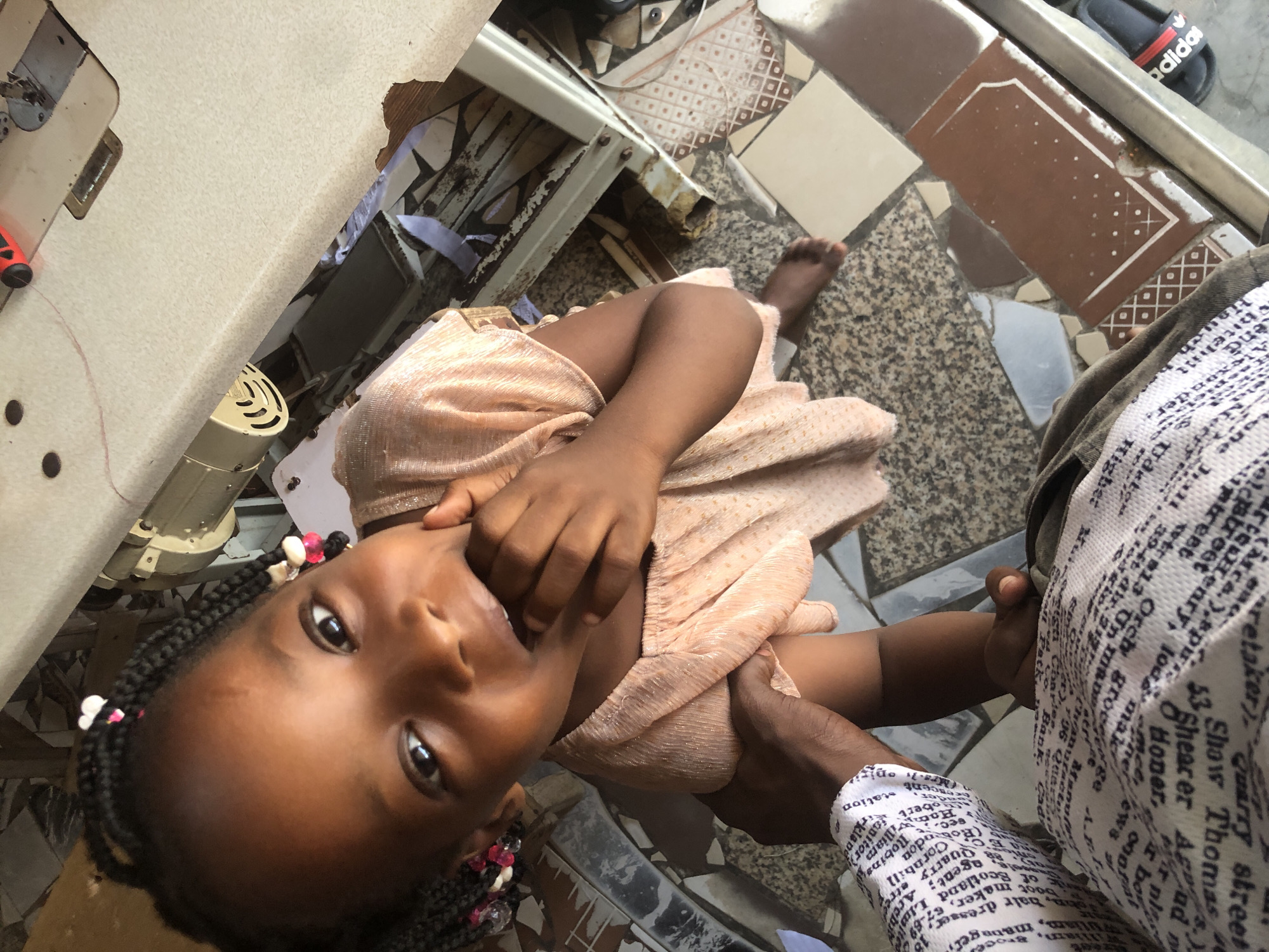 In Ado, Nigeria, an overhead shot shows a young Black girl with braided hair adorned with pink and white beads, wearing a light peach-colored dress. She is looking directly at the camera with her fingers in her mouth. A person's arm, wearing a white shirt printed with black text, is holding the girl's arm. They are standing on a floor with a mosaic of granite and tile pieces. In the background, a sewing machine is visible on a table, suggesting a domestic or workshop setting. Some pieces of fabric and debris are scattered around the sewing machine. A pair of black Adidas slides are on the floor to the right.