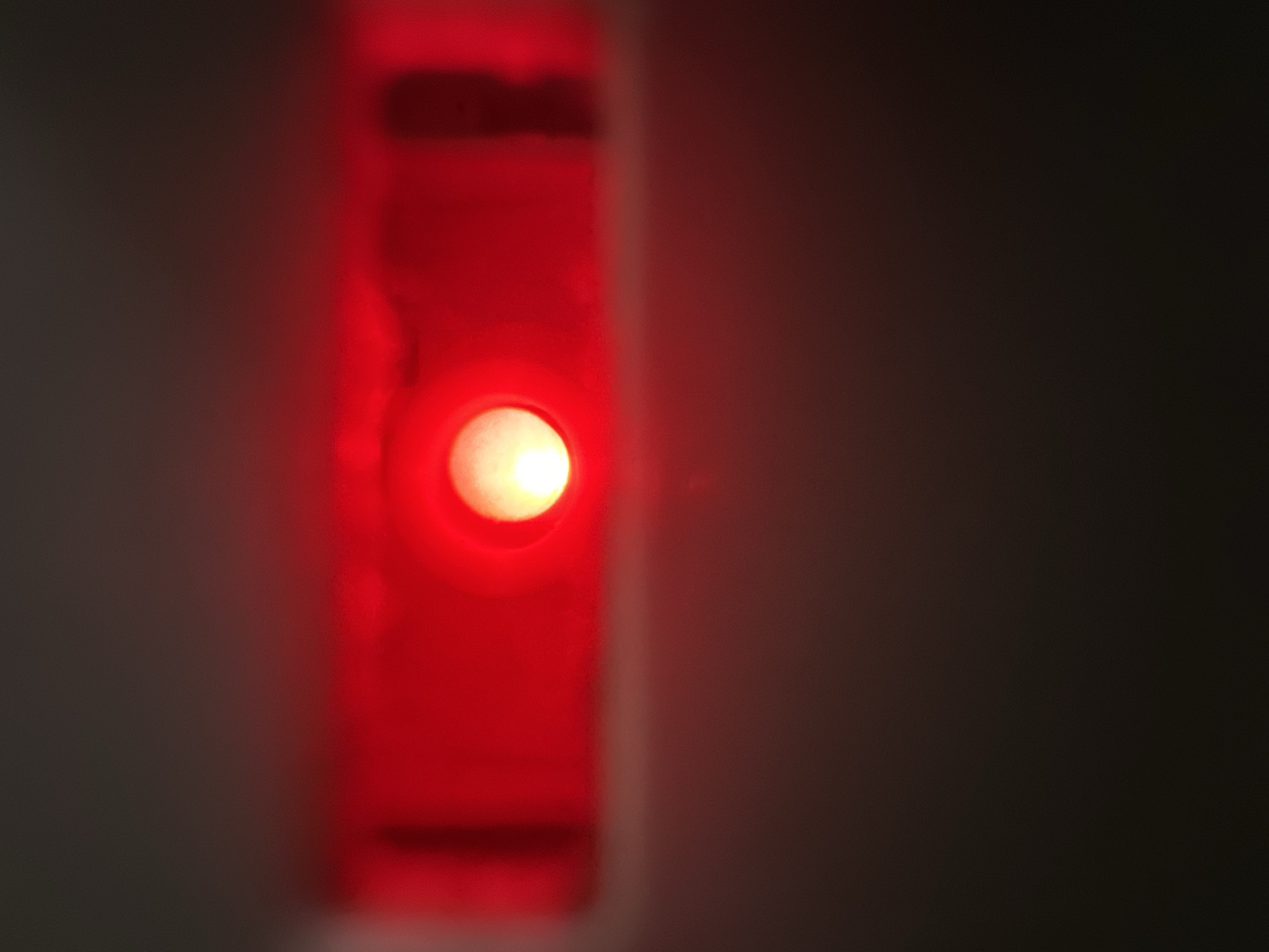 The image is a close-up, shallow depth-of-field shot of a bright red LED light illuminated within a dark, vertical rectangular opening. The light is the primary subject, casting a strong red glow that bleeds out into the surrounding area. The opening appears to be part of a device or enclosure, with the red light possibly indicating a power status, an alert, or a functional indicator. The background on the right side of the image is dark and out of focus, providing a stark contrast to the luminous red light. The left side of the image is also dark and blurry, with some indistinct shapes and textures suggesting the wall or surface surrounding the opening. There is no visible text or discernible environmental cues to indicate the specific time of day, weather, or precise location, other than the general context provided. The overall mood is abstract and focused on the intense light source.