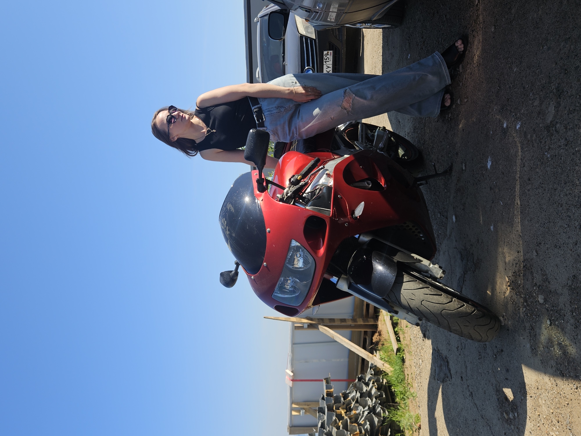 A young woman with sunglasses is leaning against a red motorcycle in an outdoor setting. She is wearing a black top and ripped blue jeans. A white and gray van is parked behind the motorcycle. The foreground features a pile of dark objects, possibly tires or debris. The sky is clear and blue. A fence and wooden supports are visible in the background. The license plate on the van says "KY 159." The location is Chaikovsky, Russia.
