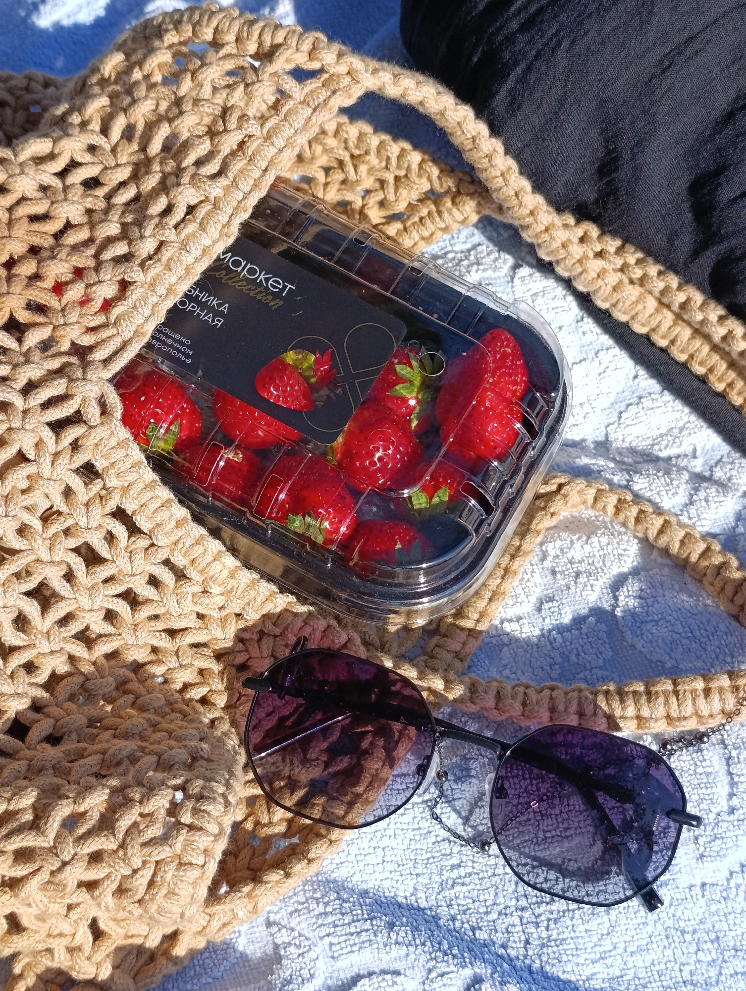 This media file shows a close-up of a straw tote bag resting on a white towel. Inside the bag, a clear plastic container filled with fresh strawberries is visible. A pair of dark-lensed sunglasses with thin metal frames and decorative chains is placed on the towel in front of the bag. The scene is bathed in bright sunlight, suggesting a sunny day, likely outdoors given the presence of a towel and sunglasses. The overall impression is one of a relaxed, summery setting, perhaps a beach or a park.
On the strawberry container, there is visible text in a dark font against a black background. Part of the text reads "маркет Collection" and below it, "ОРНАЯ". There is also some smaller text below, but it is too blurry to decipher. The strawberries appear ripe and vibrant red, with green tops. The straw bag has a distinctive macrame-like pattern. The sunglasses have a slight purple tint. The lighting creates shadows on the towel and highlights the textures of the bag and the towel. There is a dark fabric, possibly clothing, visible in the upper right corner of the image.