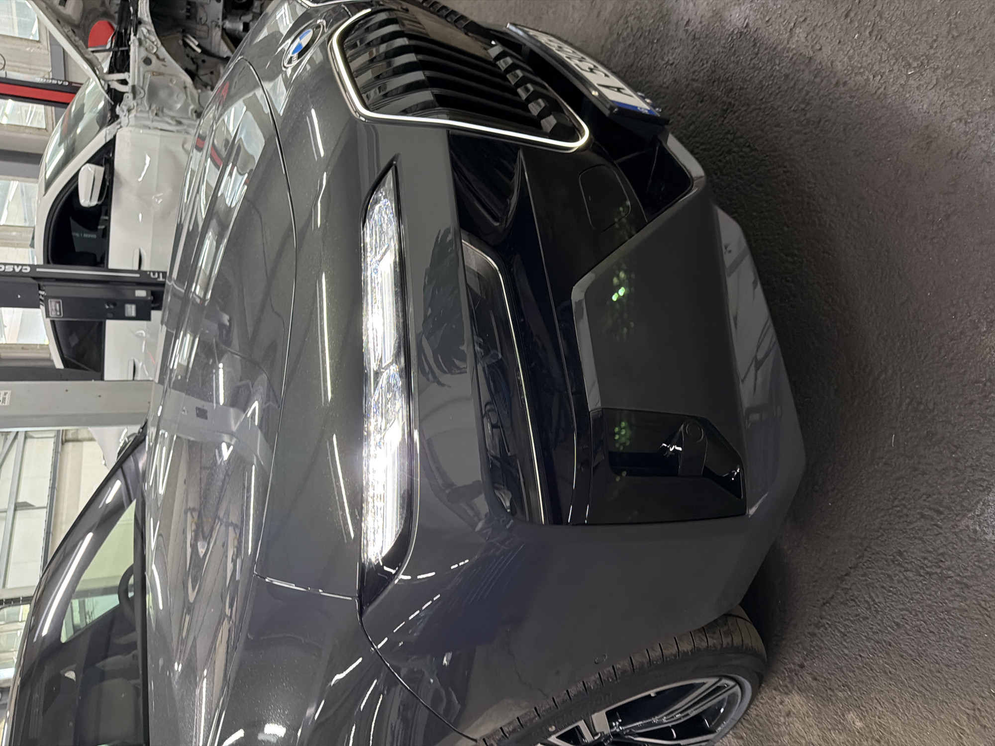 The image shows the front passenger side of a dark gray BMW sedan parked indoors on a concrete floor. The car's headlights are on, emitting a bright white light. The front grille is prominent, with vertical chrome slats outlined by an illuminated strip. A license plate is visible, partially obscured, with lettering and numbers. To the right of the BMW, a white car is partially visible, appearing to be undergoing some sort of repair or modification, with its hood open and some body panels removed. The background suggests an auto repair shop or a similar facility, with industrial lighting and equipment. There are no people visible in the image. The location is identified as Burgas, Bulgaria.