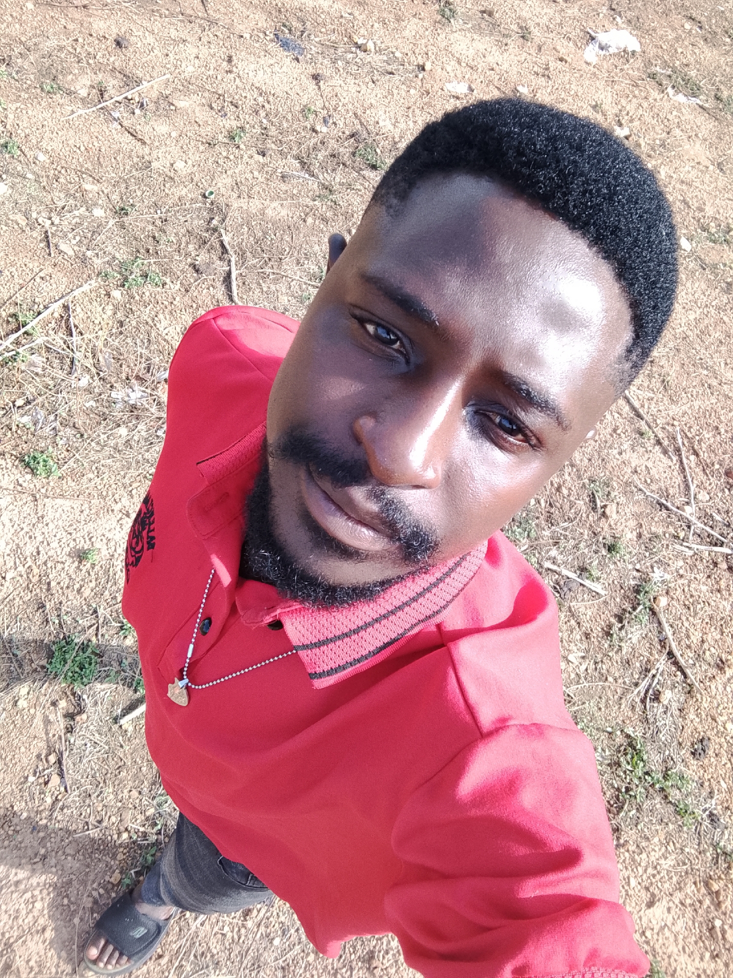 A man in a red polo shirt stands outdoors on a dusty, sandy patch of land with sparse vegetation. The man has dark skin, short black hair, a mustache, and a beard. He is wearing a necklace with a pendant. He is looking up at the camera. He is wearing gray jeans and black sandals. The image appears to be a selfie. The background consists of dry soil with small plants and debris. The location is presumed to be Akwanga, Nigeria.